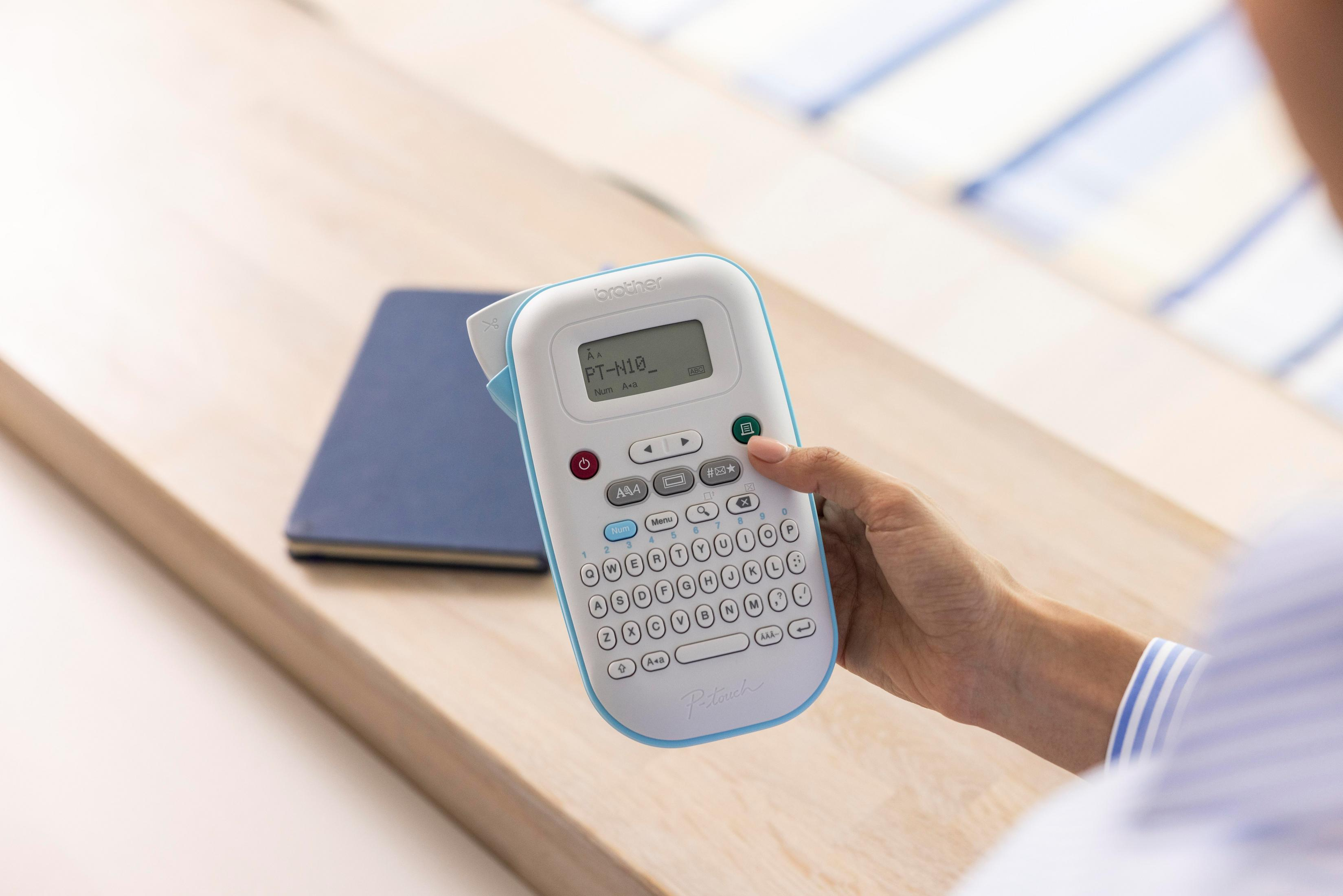 Eine Person hält einen Brother P-touch N10 Etikettendrucker auf einem Holztisch mit Notizbuch.