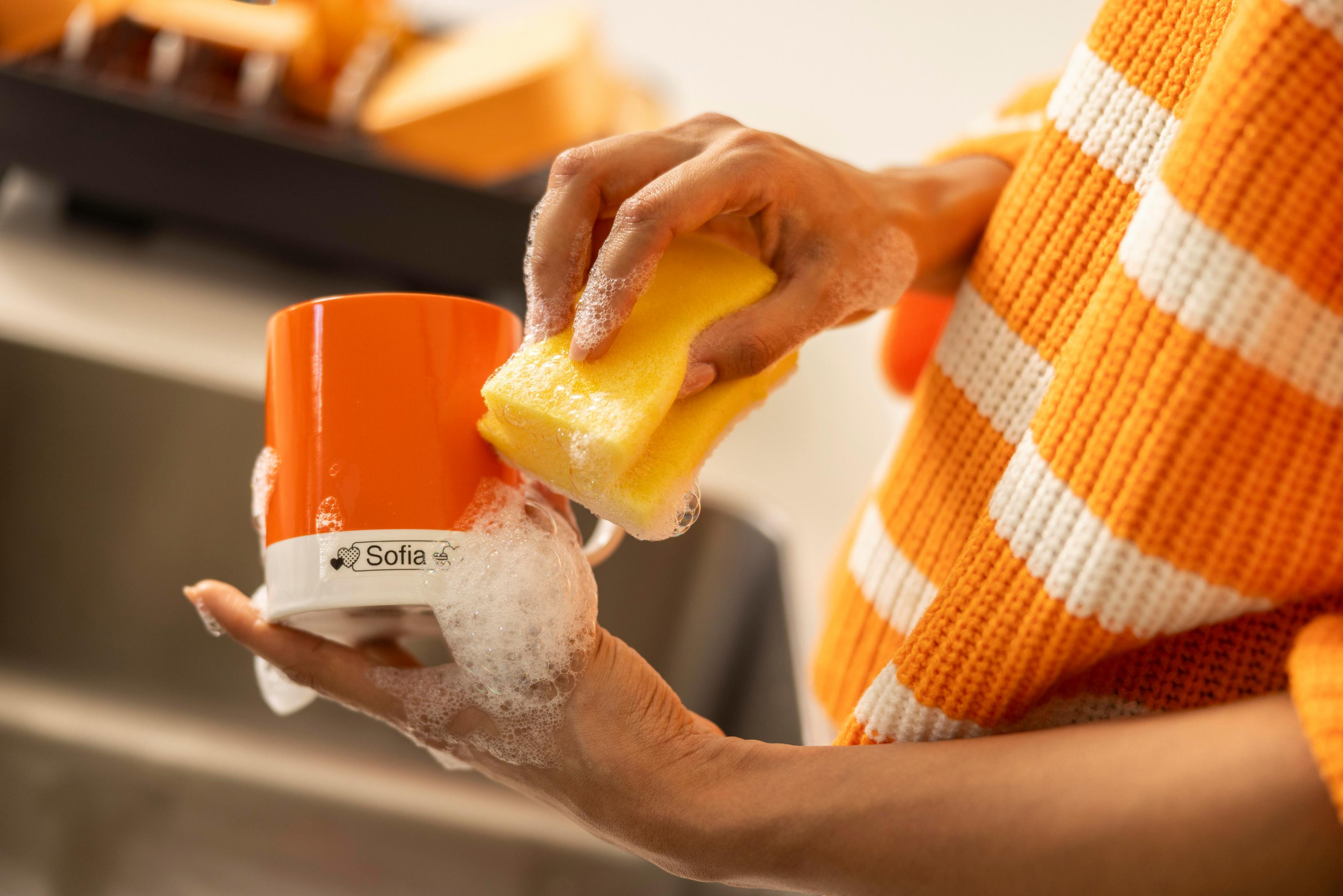 Eine Person wäscht eine orangefarbene Tasse mit einem Schwamm. Die Tasse hat den Namen 'Sofia'.