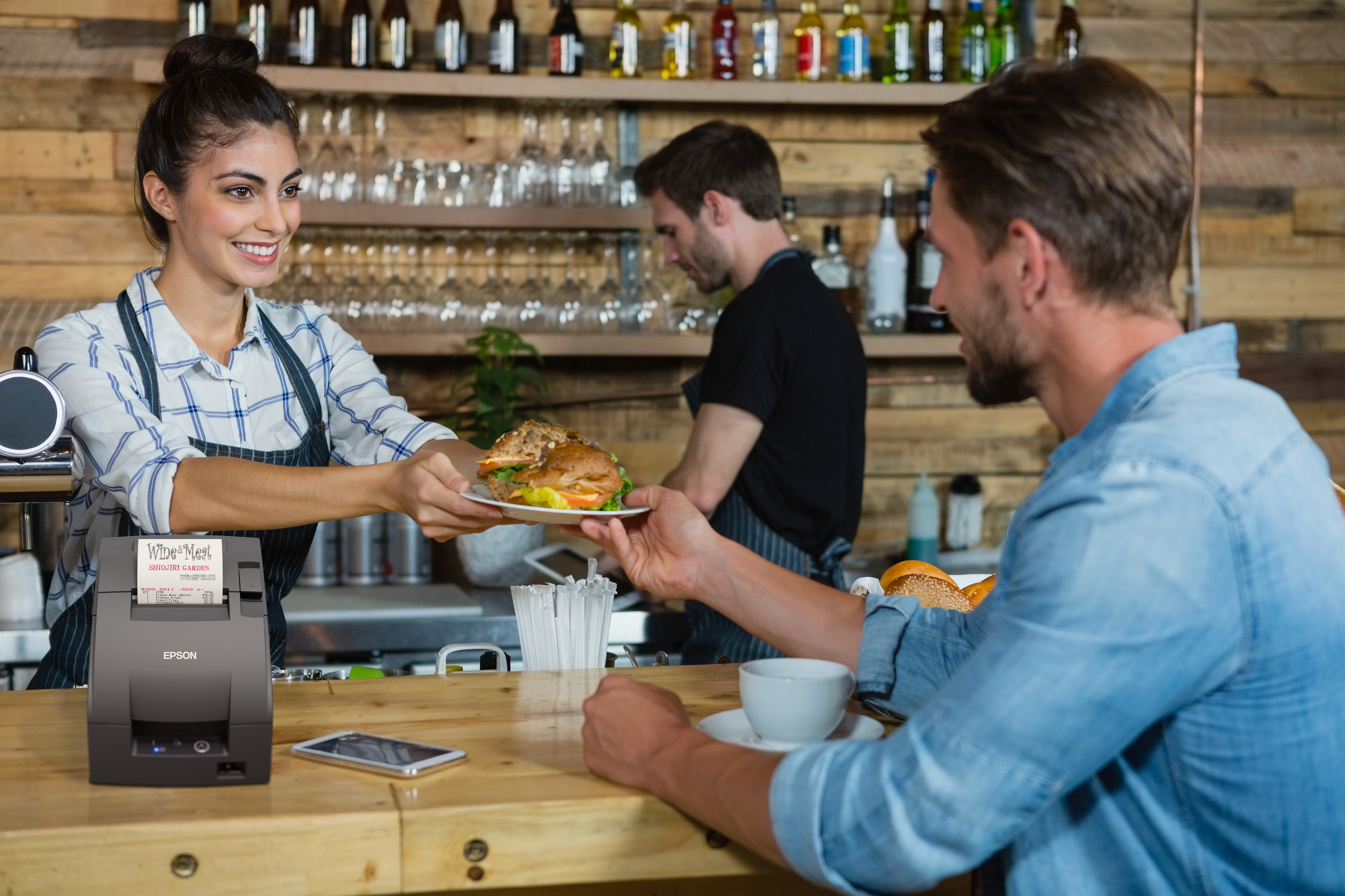 Una cameriera consegna un piatto a un cliente in un bar, con una stampante per ricevute Epson sul bancone.