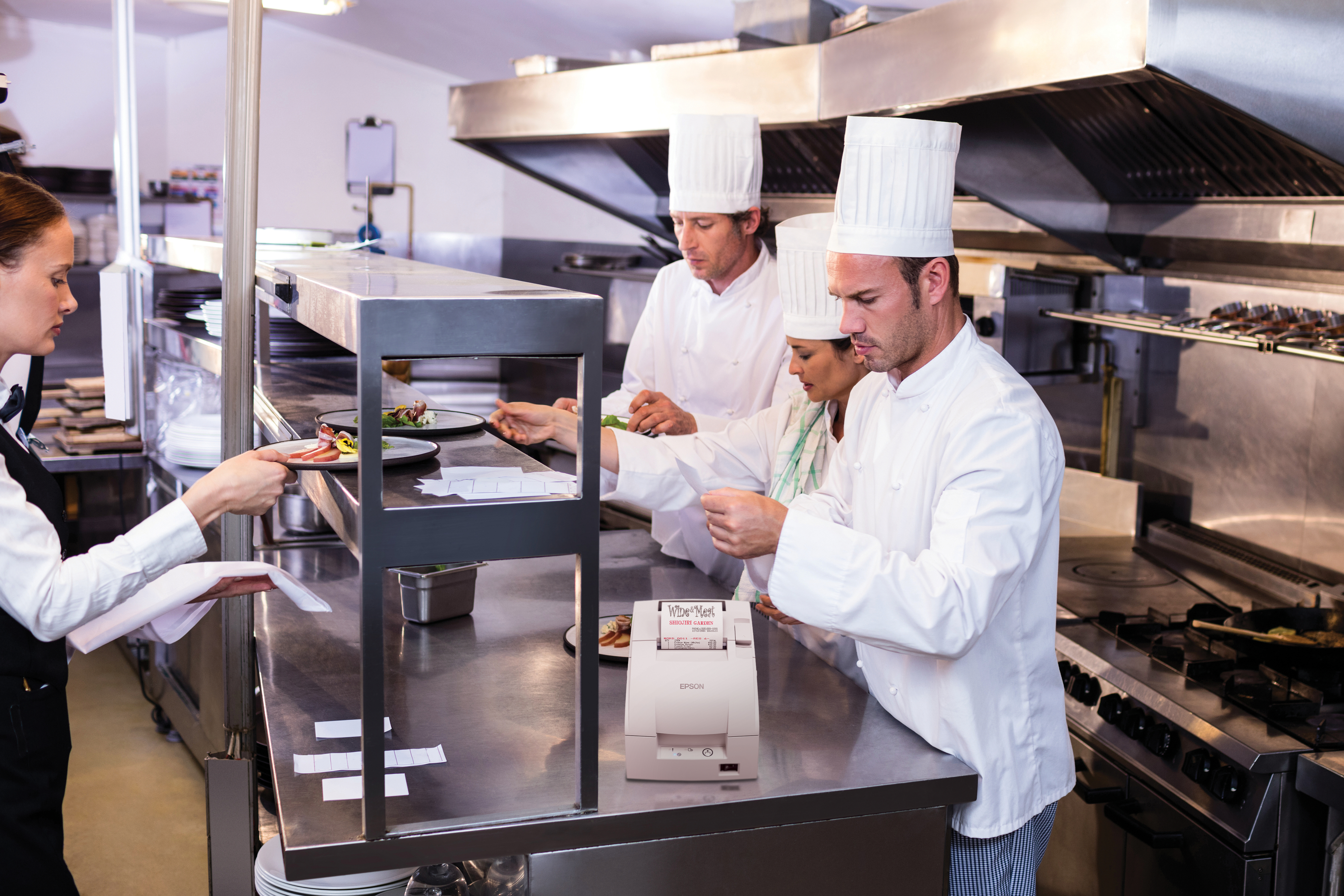 Chef in uniforme bianca servono cibo in una cucina commerciale, con una stampante Epson sul bancone.