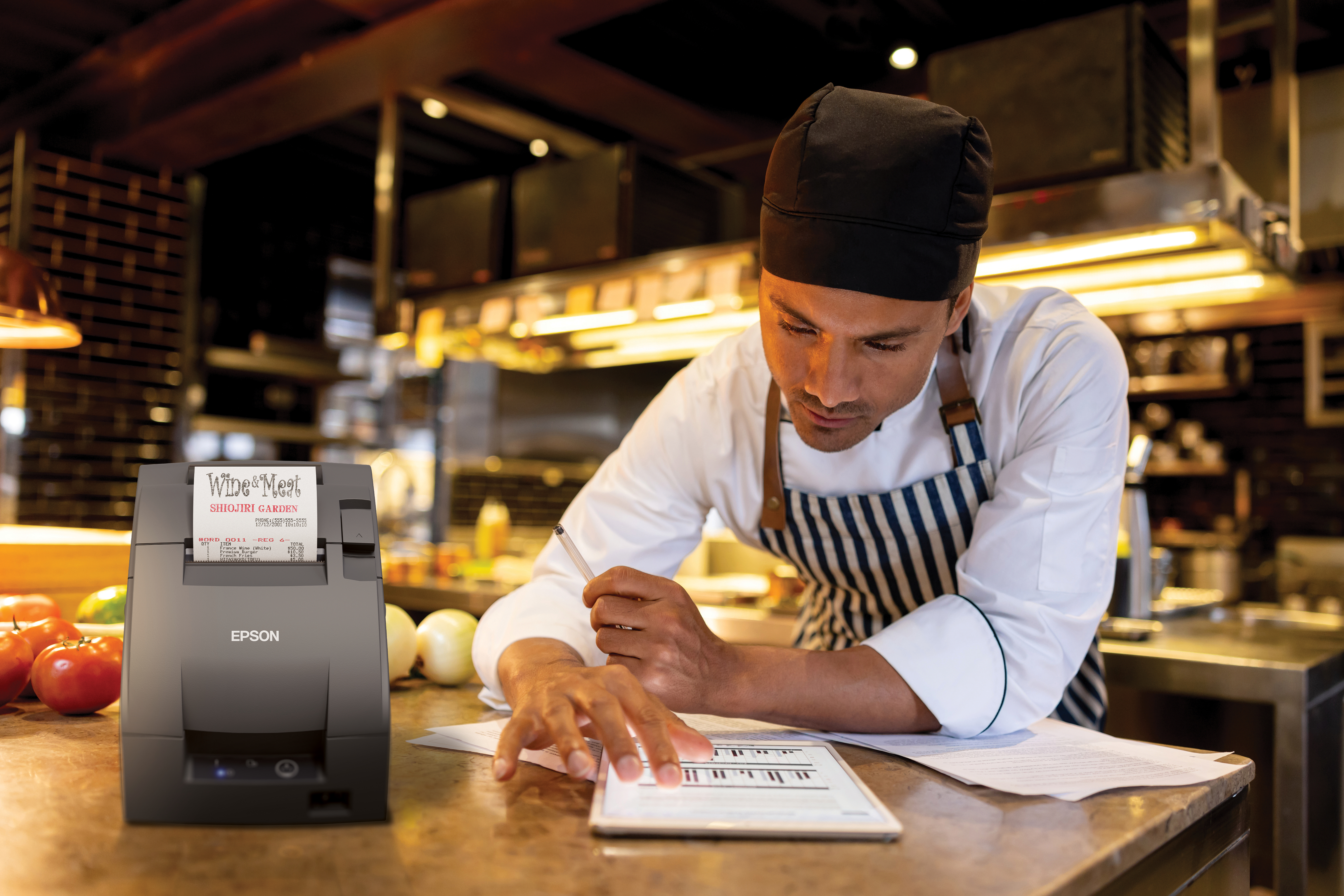 Chef in uniforme controlla un tablet accanto a una stampante Epson. Prodotti e documenti sono sul bancone.