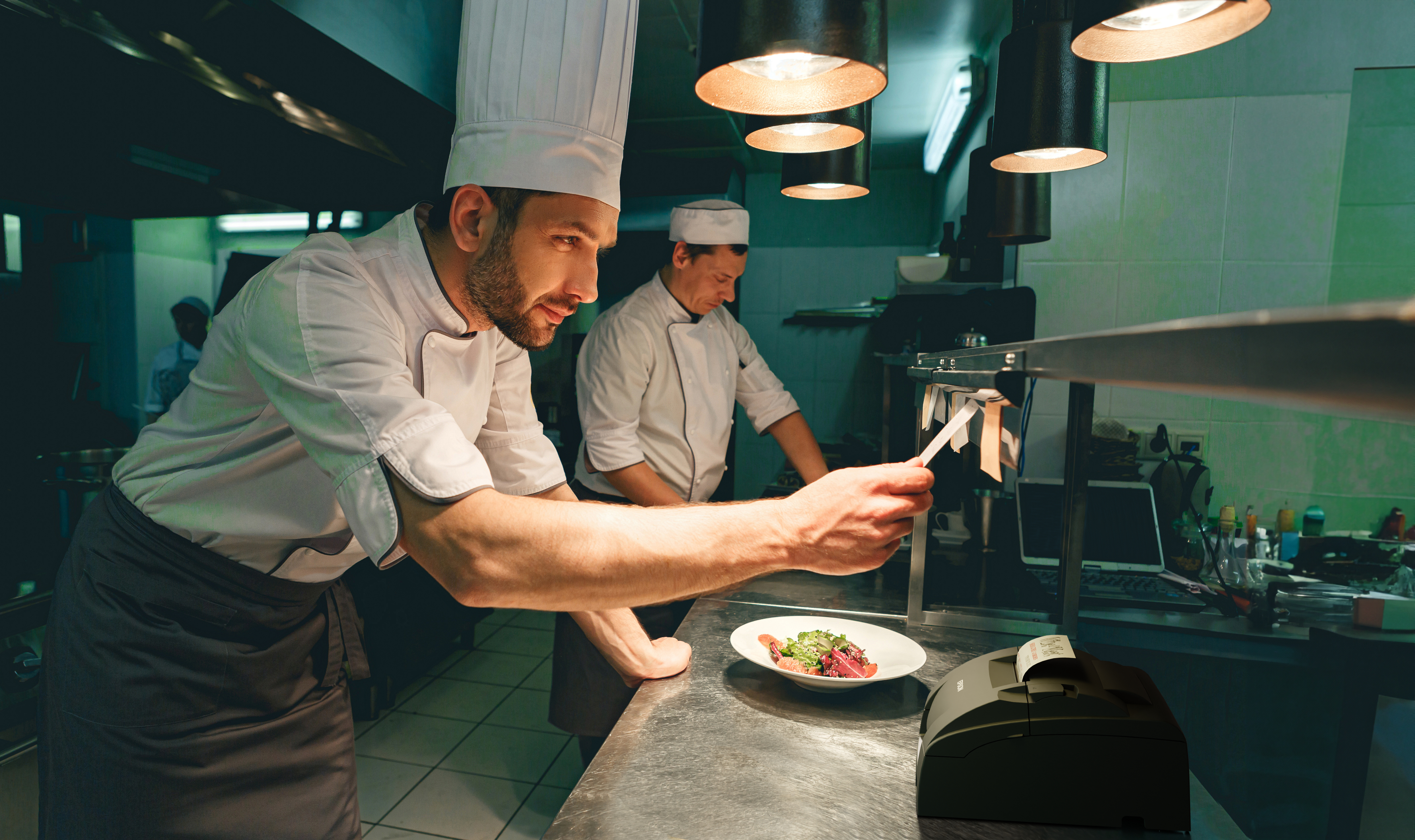 Chef che impiattano cibo in una cucina commerciale con una stampante Epson su un bancone in acciaio inossidabile.