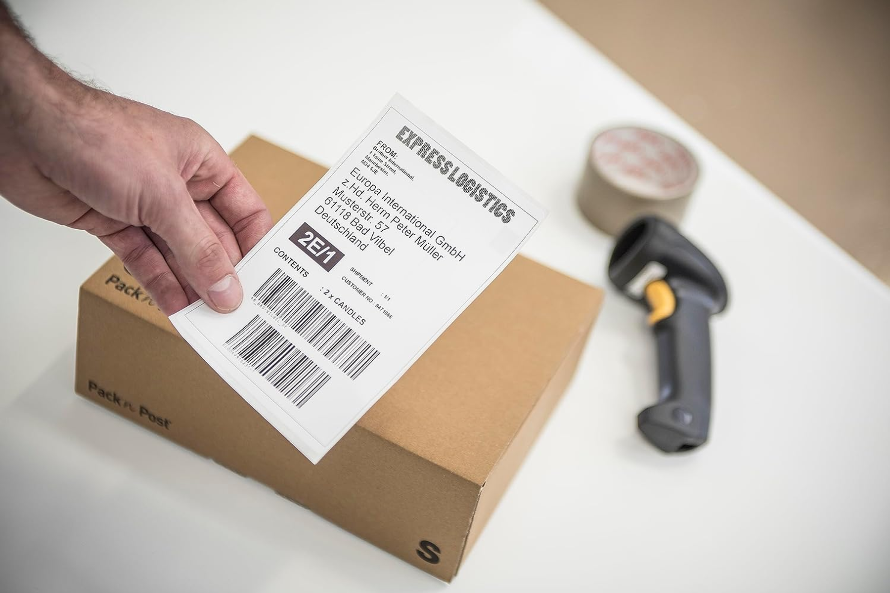 Ręka trzyma etykietę logistyki ekspresowej nad pudełkiem obok skanera kodów kreskowych i taśmy.