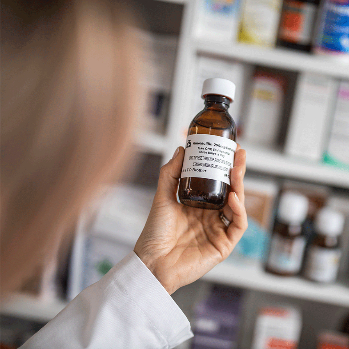 Eine Person hält eine Medikamentenflasche in einer Apotheke. Im Hintergrund sind Regale mit verschiedenen Medikamenten.