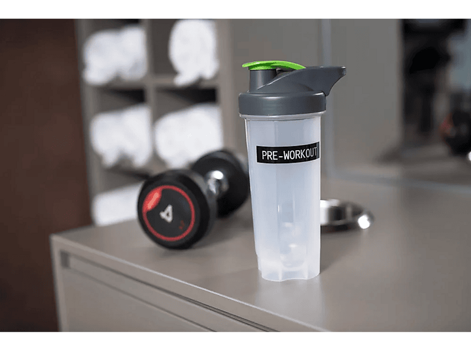 Eine Shakerflasche mit der Aufschrift "PRE-WORKOUT", eine Hantel und Handtücher stehen auf einer grauen Oberfläche.