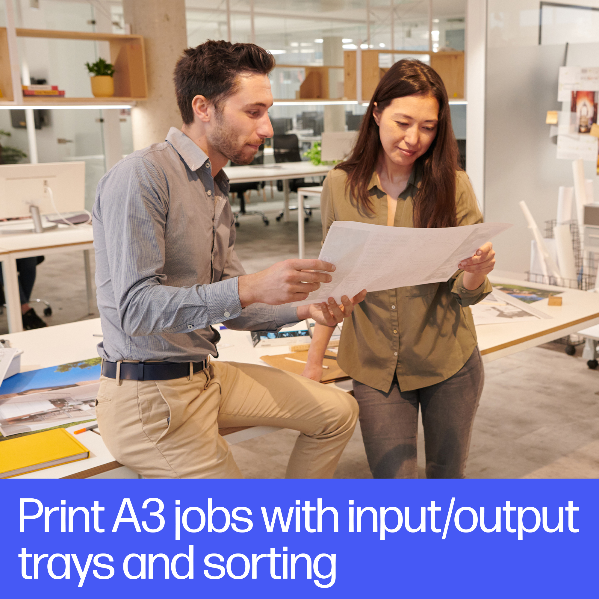 Dwie osoby przeglądają duży dokument w jasnym biurze. Na pierwszym planie znajduje się tekst "Print A3 jobs..."