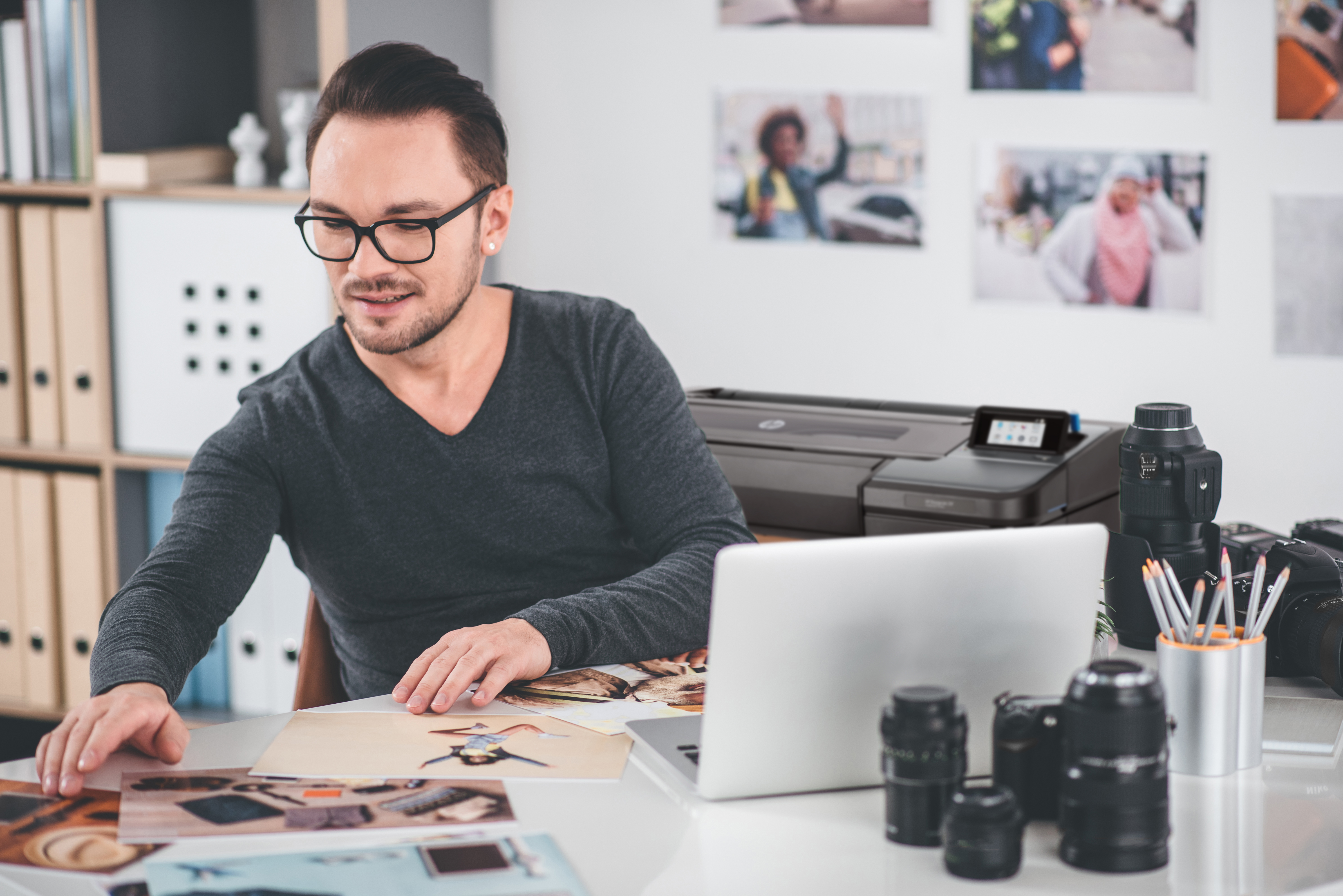 Mężczyzna w okularach siedzi przy biurku z laptopem i drukarką w tle, otoczony zdjęciami i sprzętem fotograficznym.