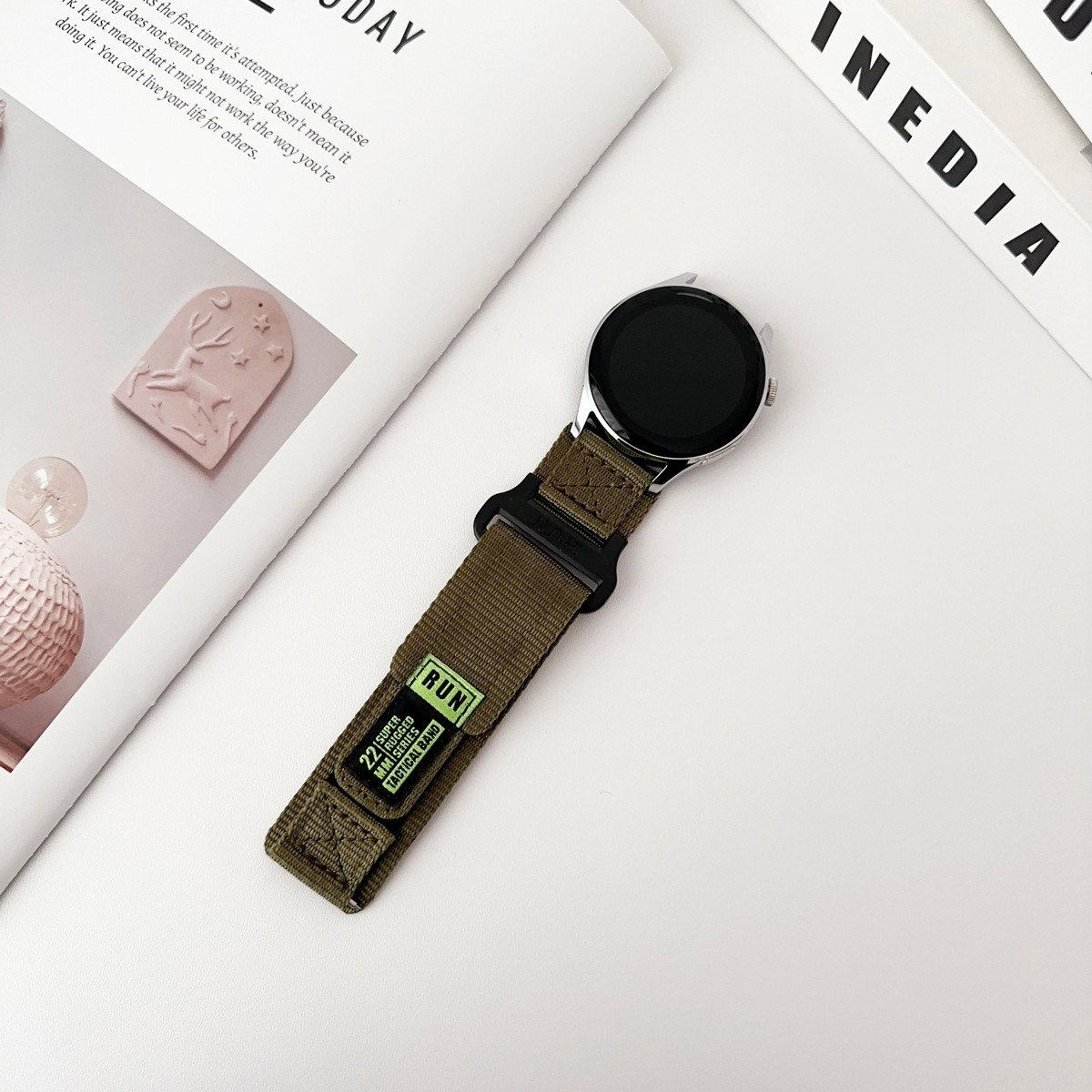 Eine Uhr mit grünem Armband liegt auf einer weißen Oberfläche mit einer Zeitschrift.