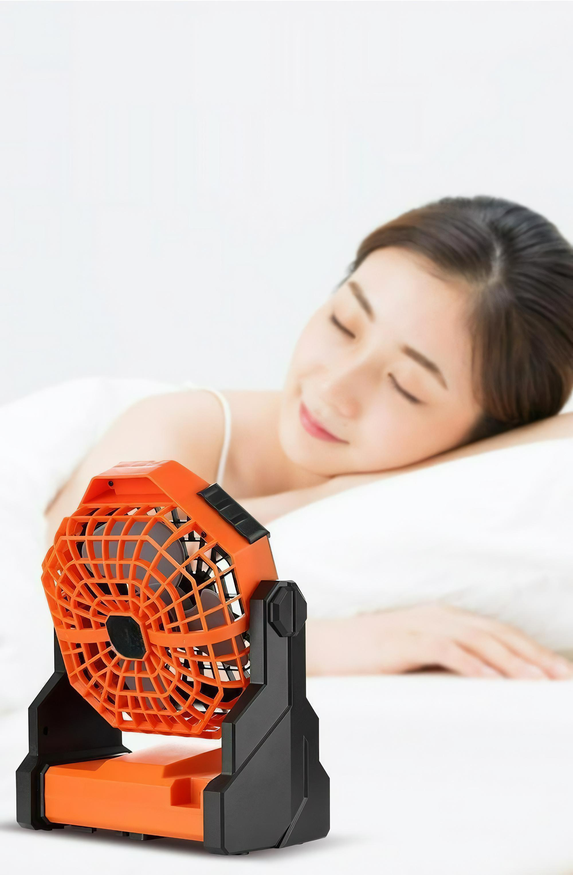 Ein orangefarbener und schwarzer Ventilator steht auf einem Bett. Im Hintergrund schläft eine Frau.