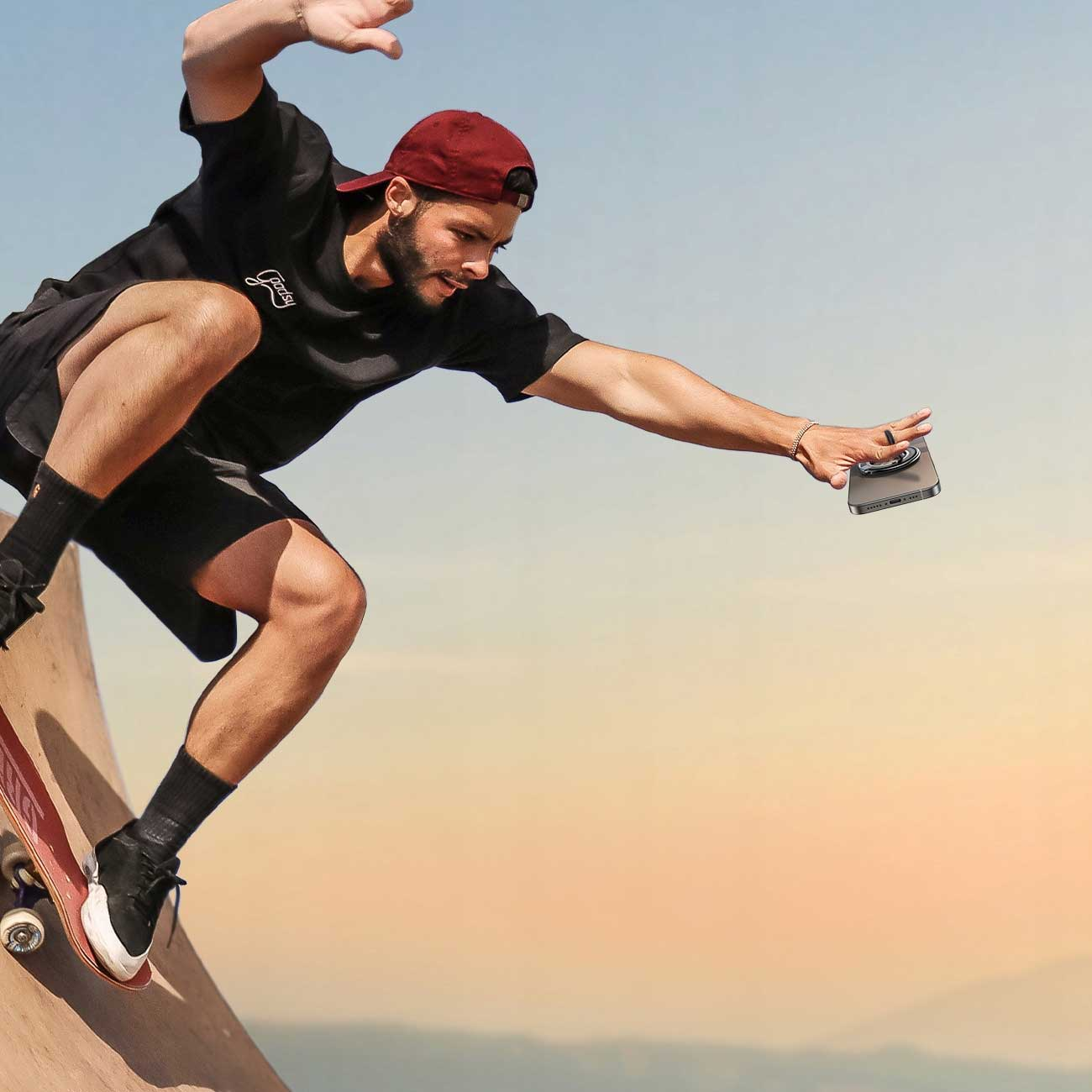 Mężczyzna na deskorolce wyciąga rękę, by złapać telefon. Tłem jest błękitne niebo.