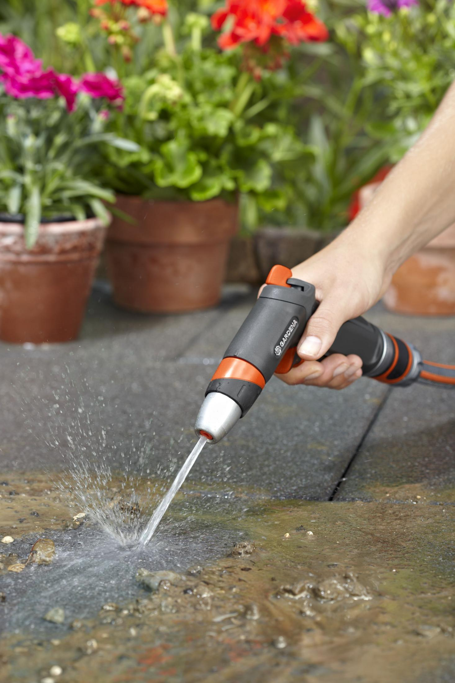 Hände halten eine GARDENA-Wasserpistole, die Wasser auf Steinfliesen sprüht, mit Blumen in Töpfen.