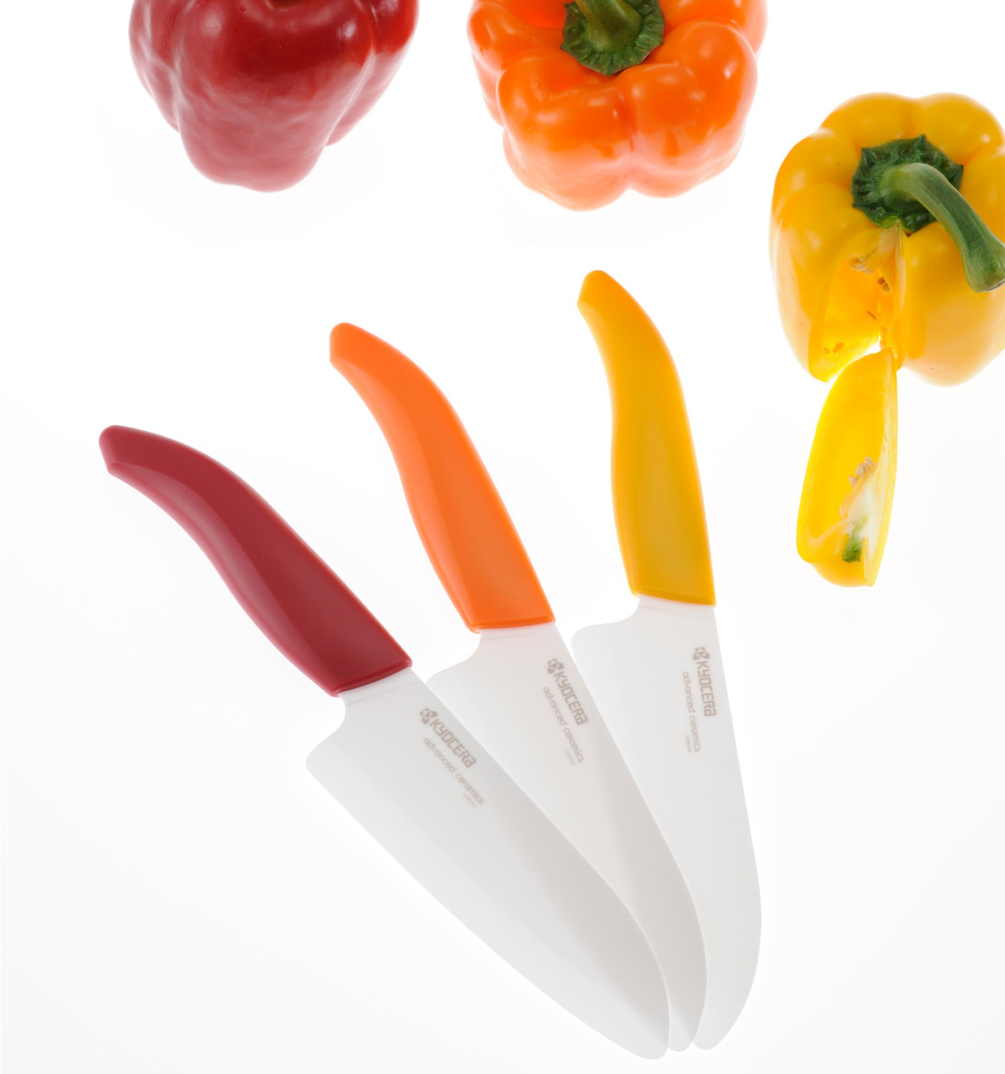 Drei Keramikmesser mit roten, orangefarbenen und gelben Griffen mit Paprika im Hintergrund.