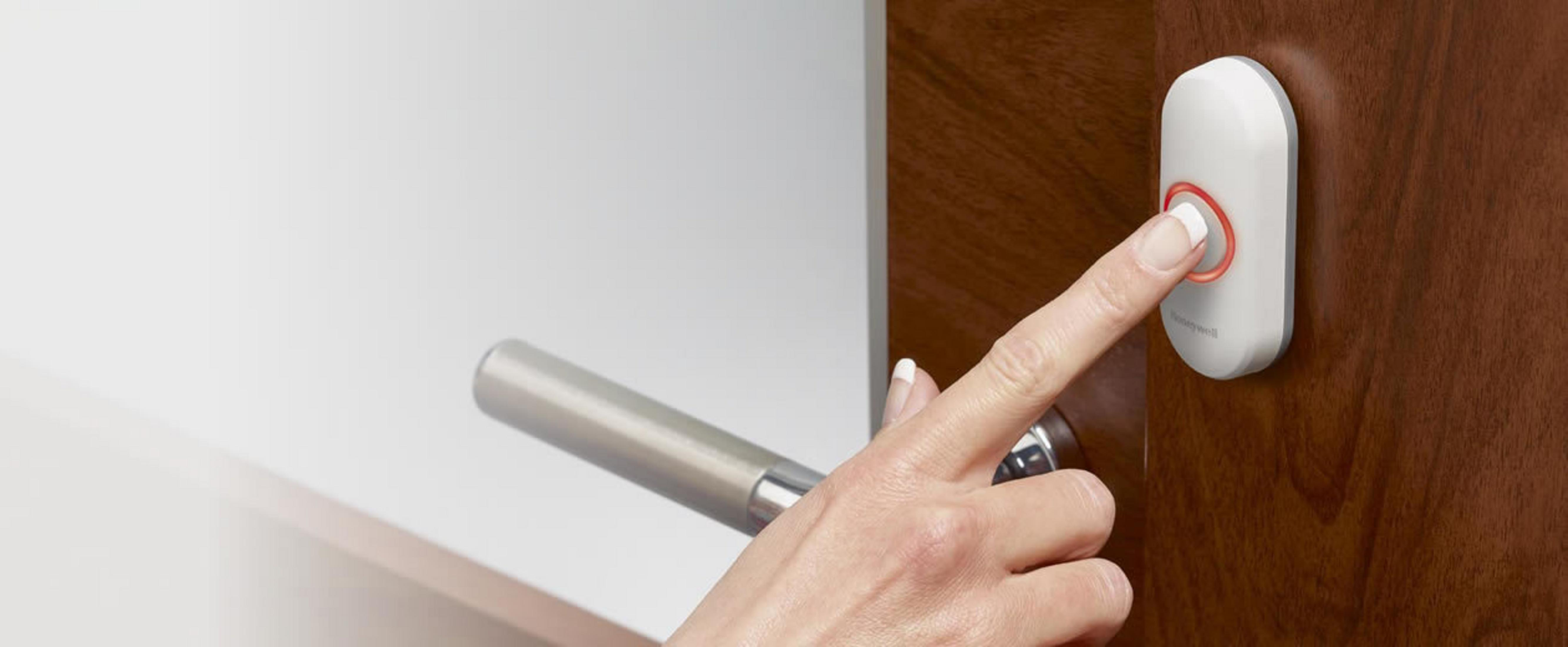 Eine Hand drückt eine weiße Türklingel mit einem roten Ring an einer Holztür. Die Tür hat einen Metallgriff.