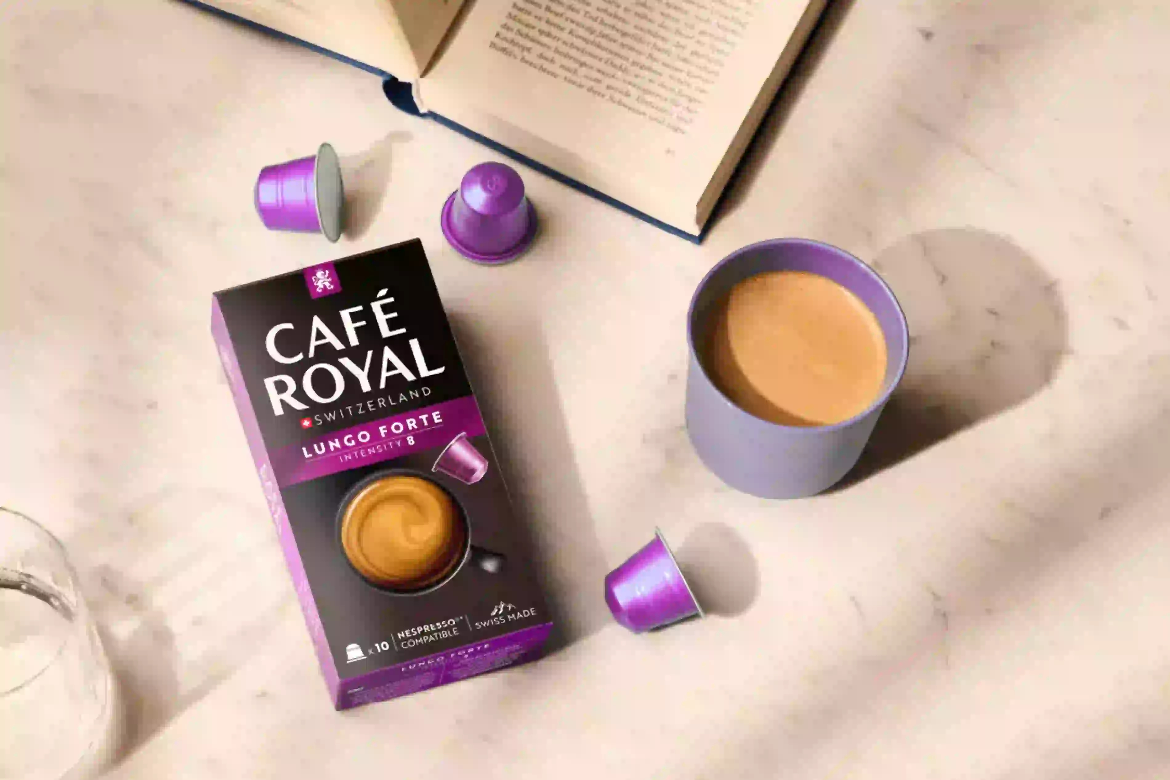 Eine Café Royal Lungo Forte Schachtel, Kapseln und eine Tasse Kaffee auf einer Marmoroberfläche mit einem Buch.