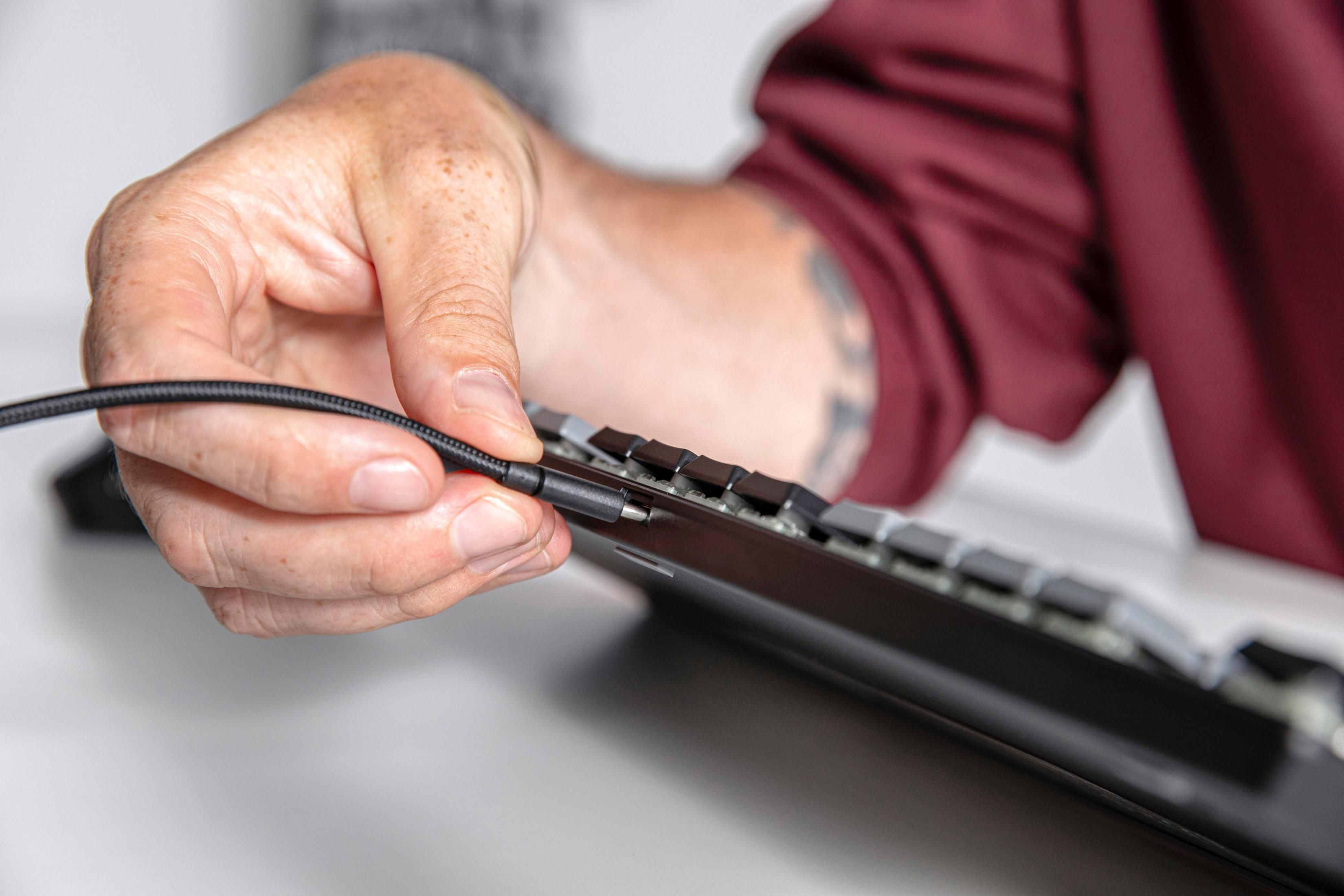 Eine Hand steckt ein Kabel in eine schwarze Computertastatur auf einem weißen Tisch.
