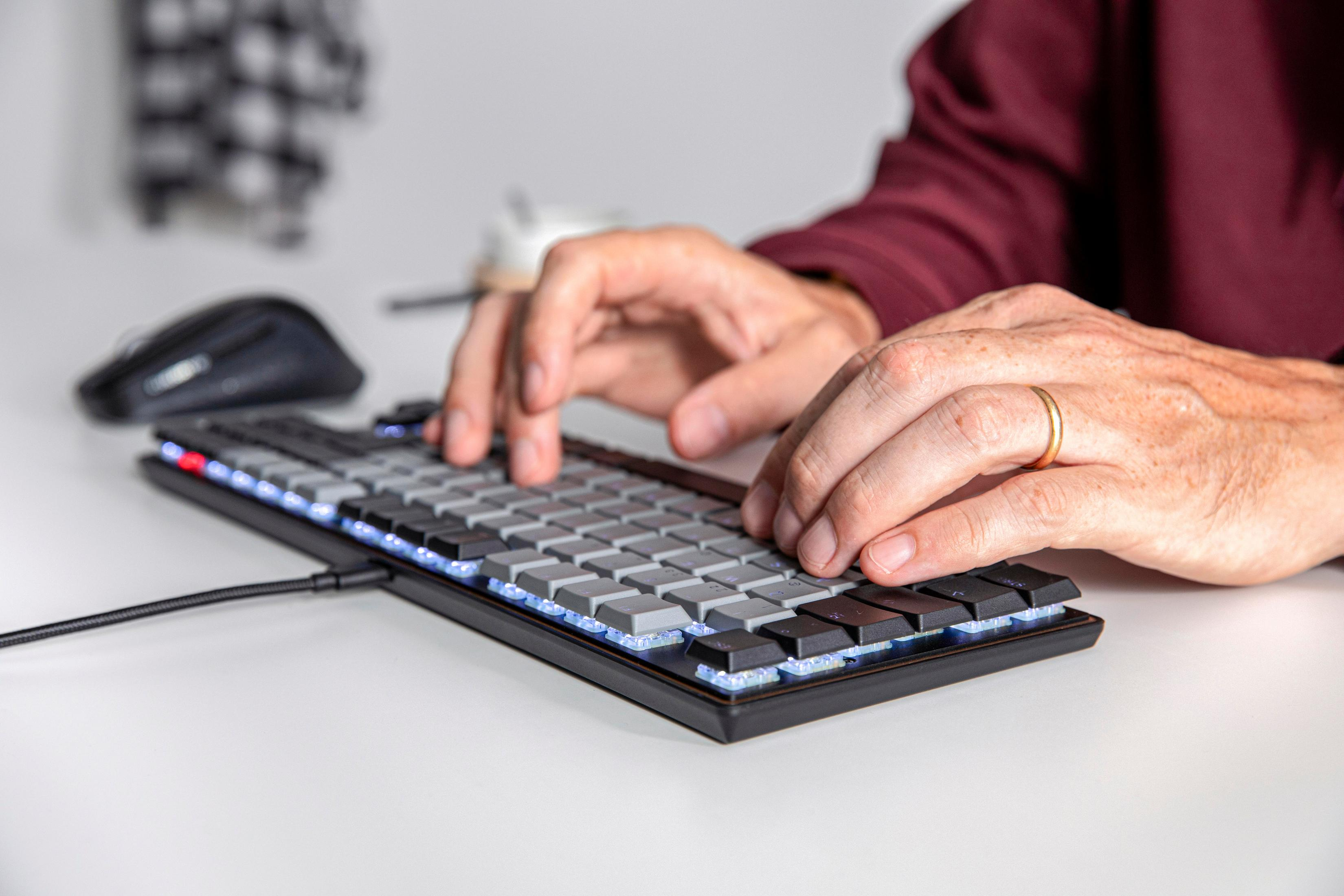 Eine Person tippt auf einer schwarzen und grauen Computertastatur auf einem weißen Tisch.