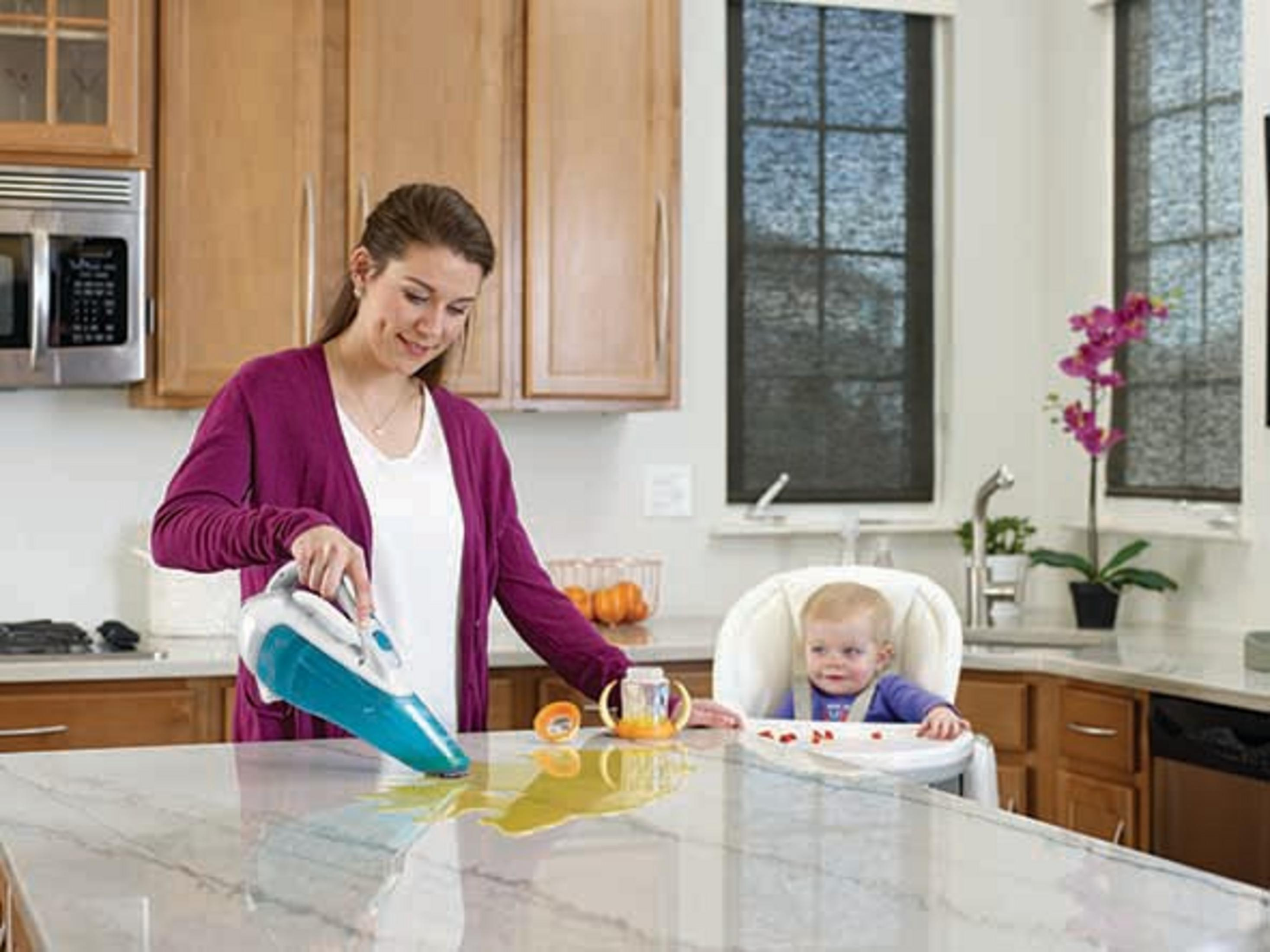 Une femme utilise un aspirateur à main sur un comptoir de cuisine avec du liquide renversé et un bébé dans une chaise haute.