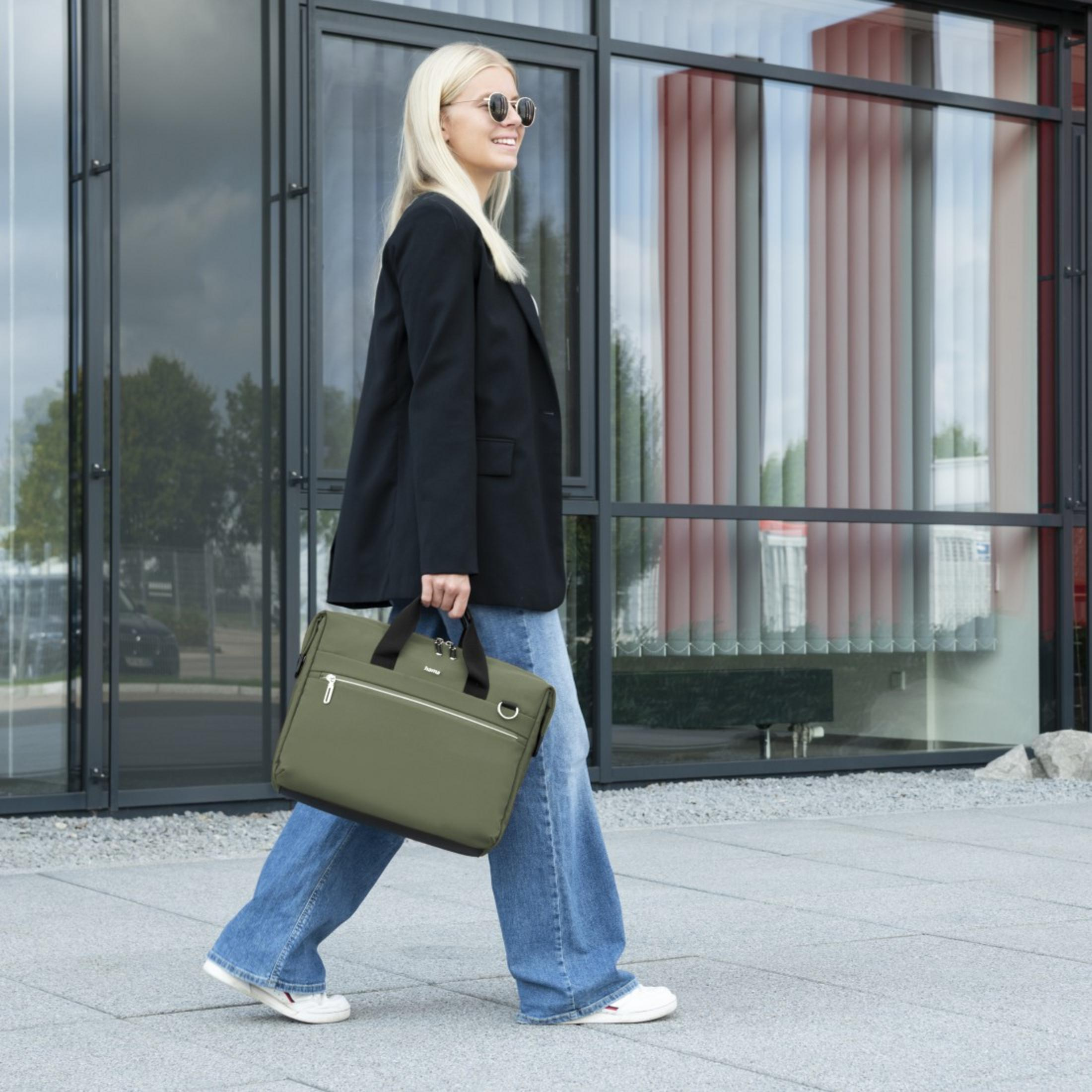 Blondynka w czarnej marynarce i niebieskich dżinsach idzie na zewnątrz trzymając oliwkową torbę na laptopa.