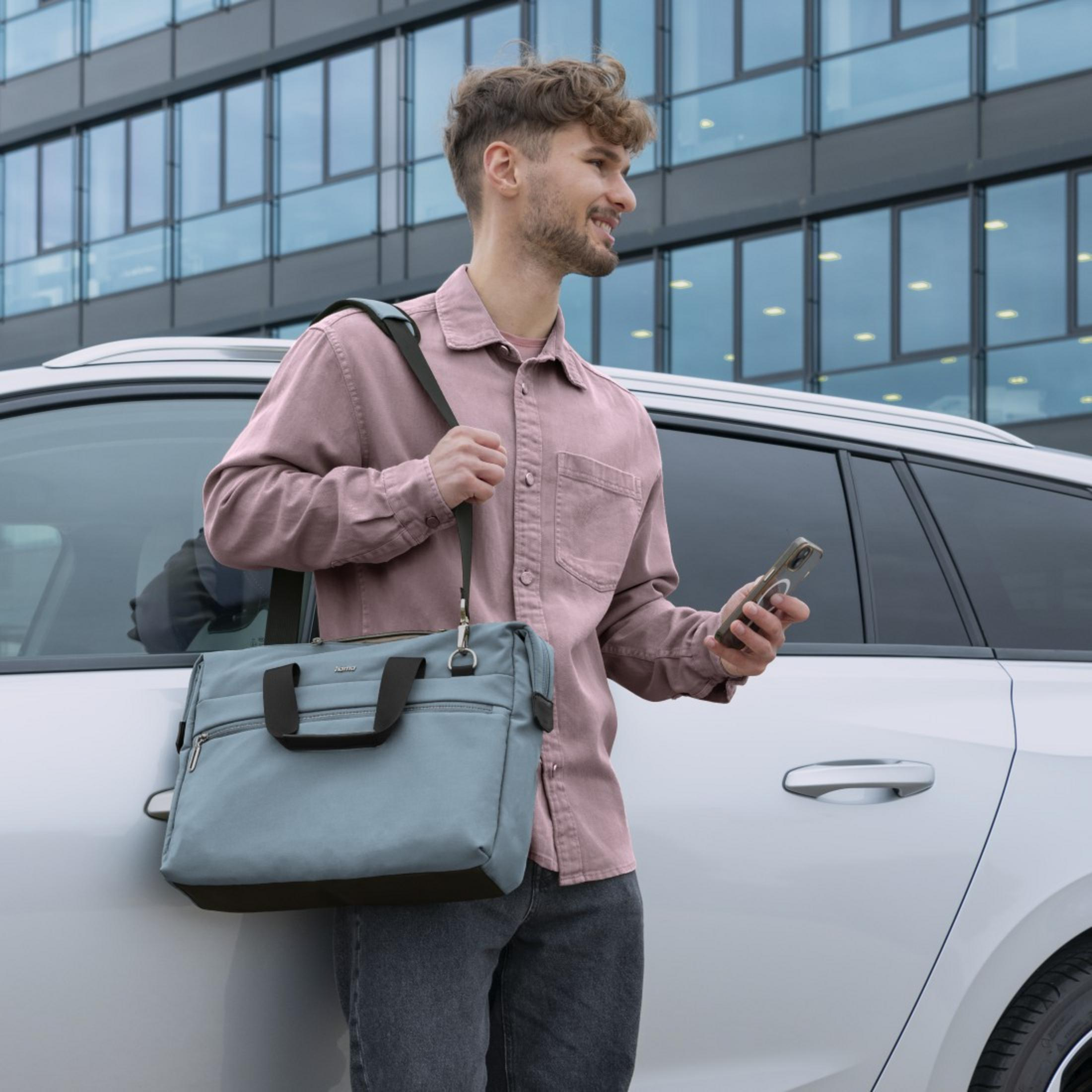 Mężczyzna z jasnoniebieską torbą, różową koszulą i telefonem stoi w pobliżu białego samochodu i budynku.