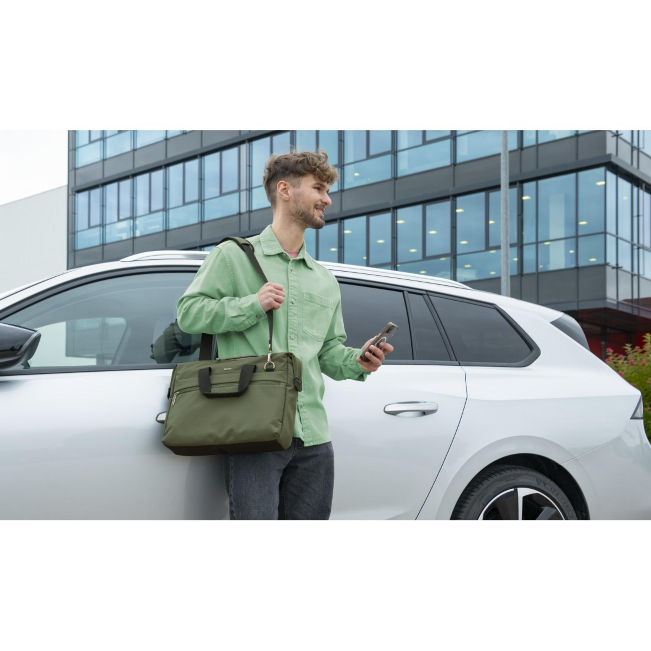 Mężczyzna w zielonej koszuli trzyma oliwkową torbę na laptopa i telefon obok srebrnego samochodu, w tle budynek.