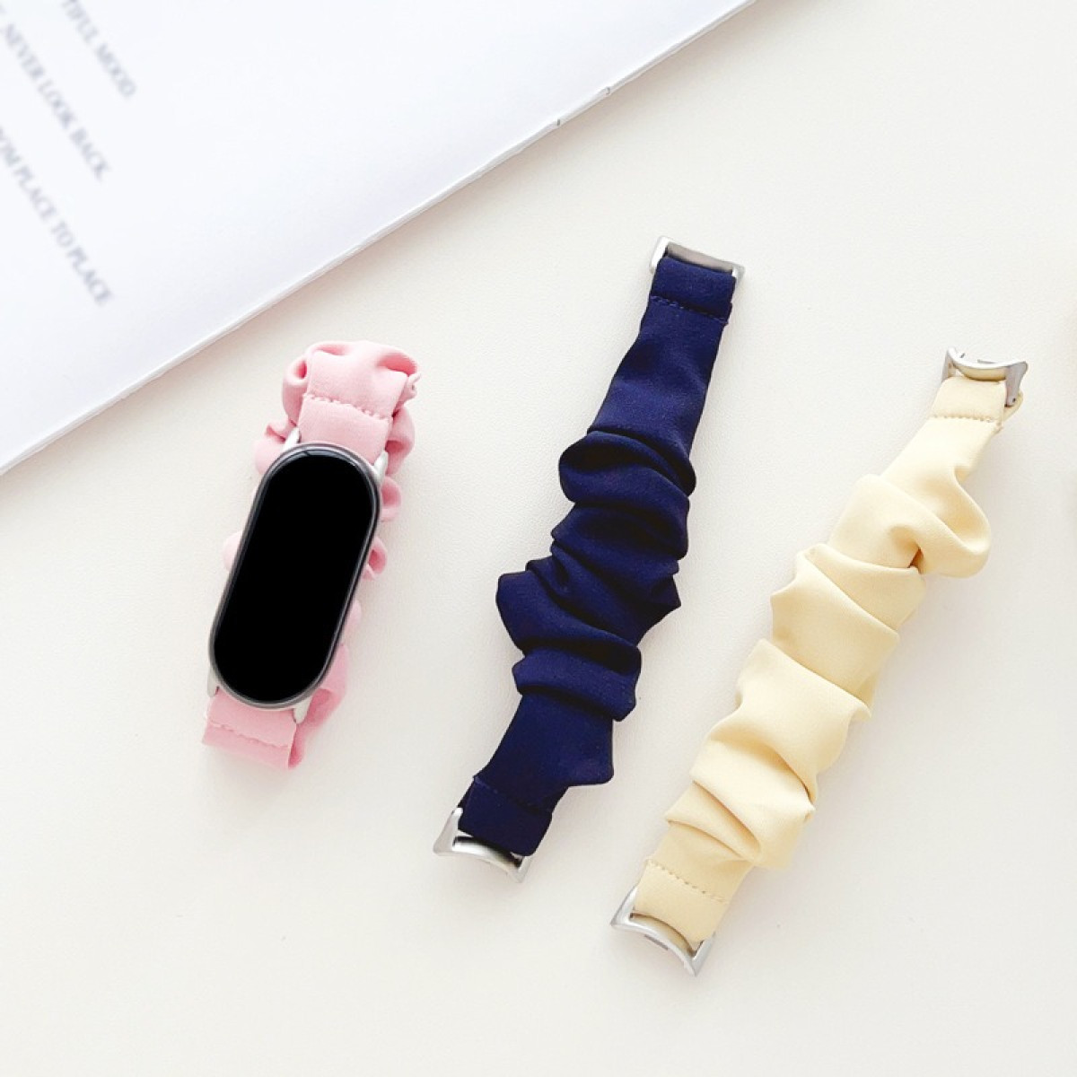 Tre cinturini per orologio scrunchie, rosa, blu scuro e beige, con uno smartwatch.