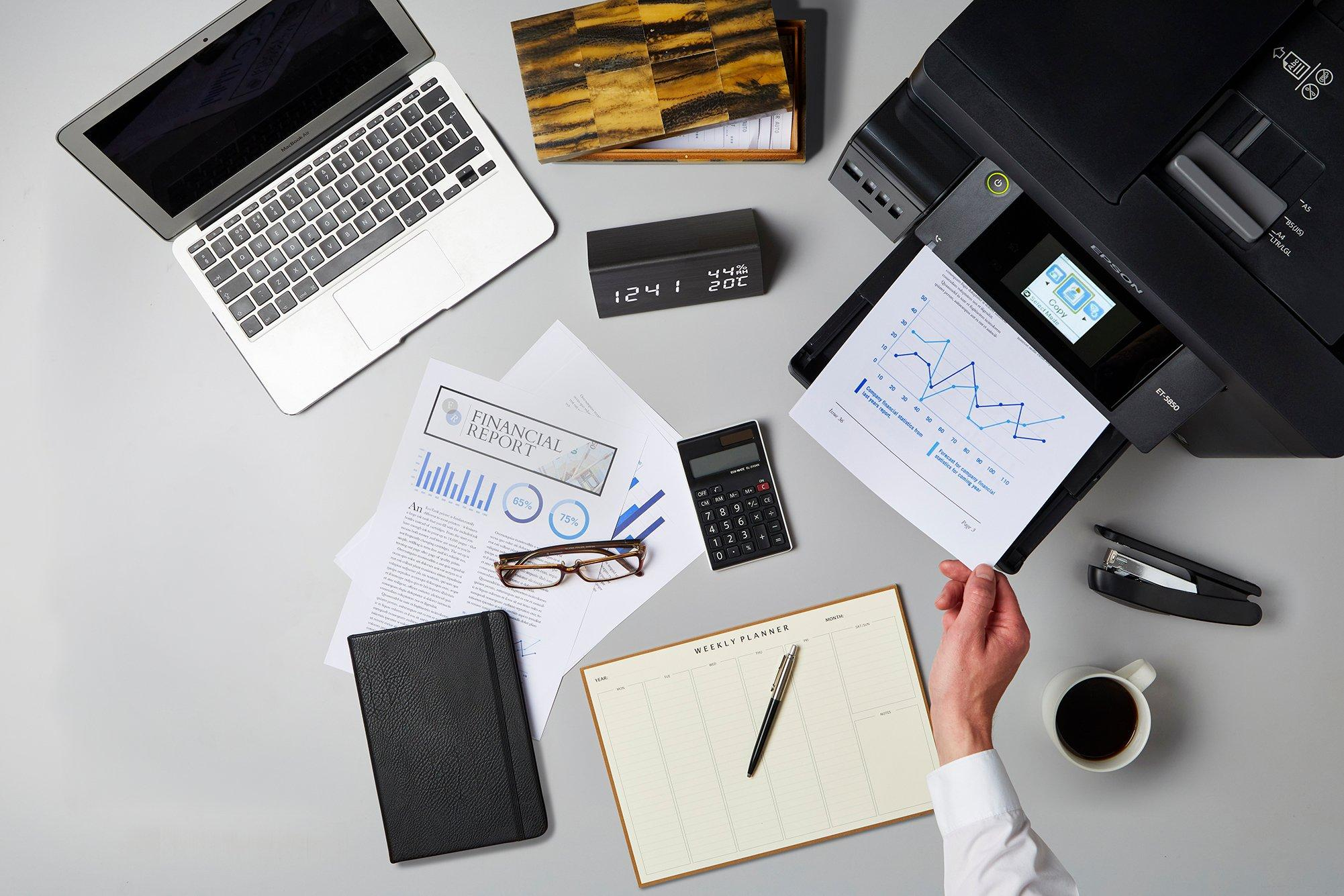 Zdjęcie z góry biurka z laptopem, drukarką, papierami, planerem, kalkulatorem, okularami i filiżanką kawy.