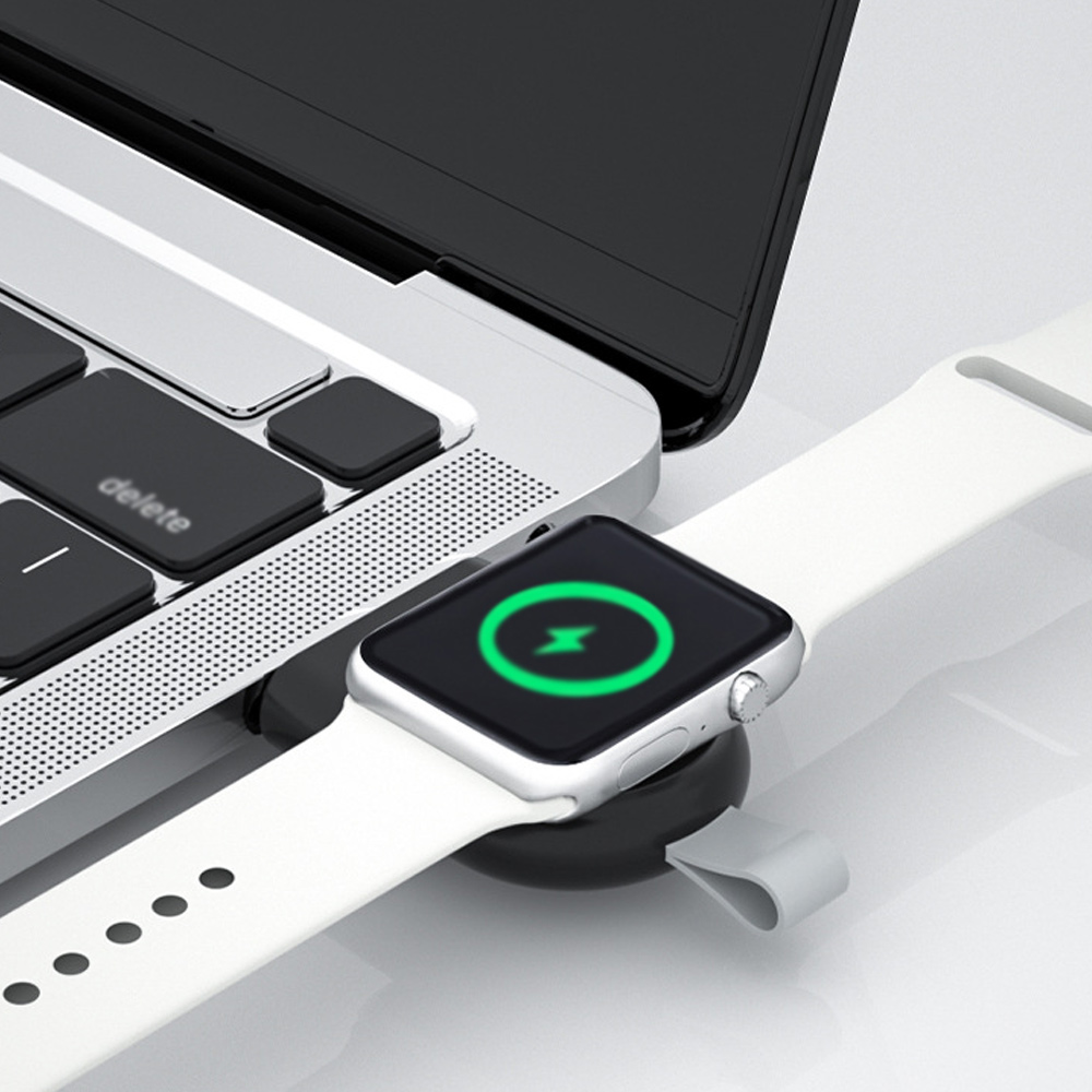 Eine silberne Apple Watch mit einem weißen Armband wird auf einem schwarzen Magnetpad neben einem Laptop aufgeladen.