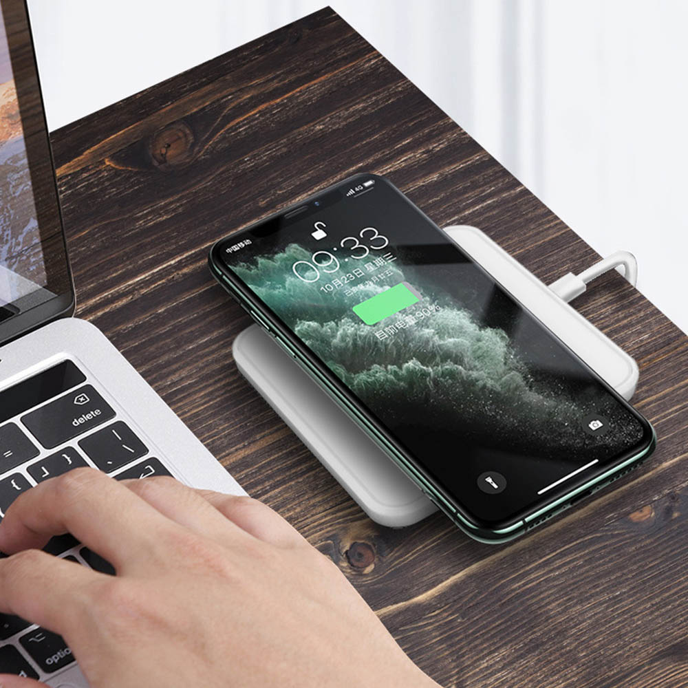 Das Bild zeigt ein Telefon, das auf einem weißen kabellosen Ladegerät neben einem Laptop aufgeladen wird.