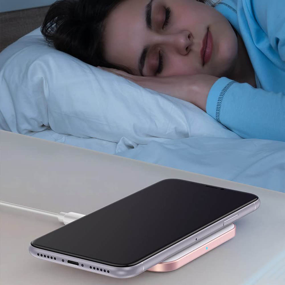 Das Bild zeigt ein Telefon, das auf einem rosafarbenen und weißen kabellosen Ladegerät auf einem Nachttisch aufgeladen wird.