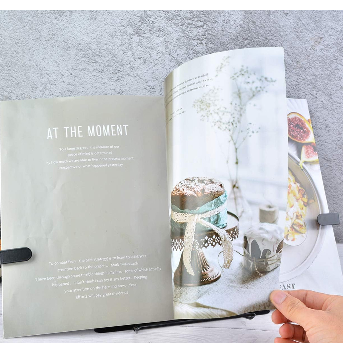 Ein Buch ist auf einem Ständer geöffnet. Die Seiten zeigen Text und Fotos von Essen, Blumen und Desserts auf einer strukturierten Oberfläche.