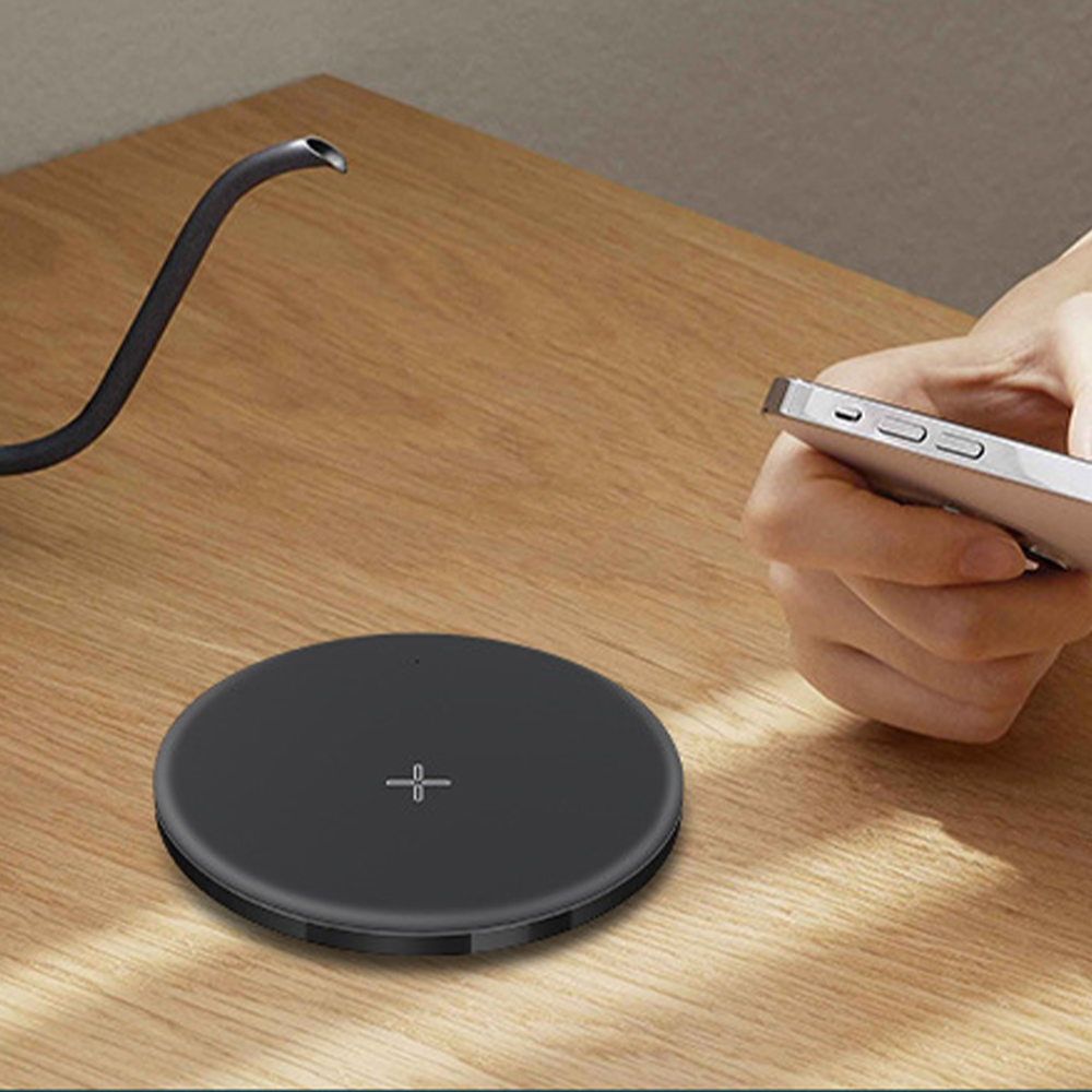Ein schwarzes, kabelloses Ladepad auf einem Holztisch mit einer Hand, die ein weißes Telefon im Hintergrund hält.