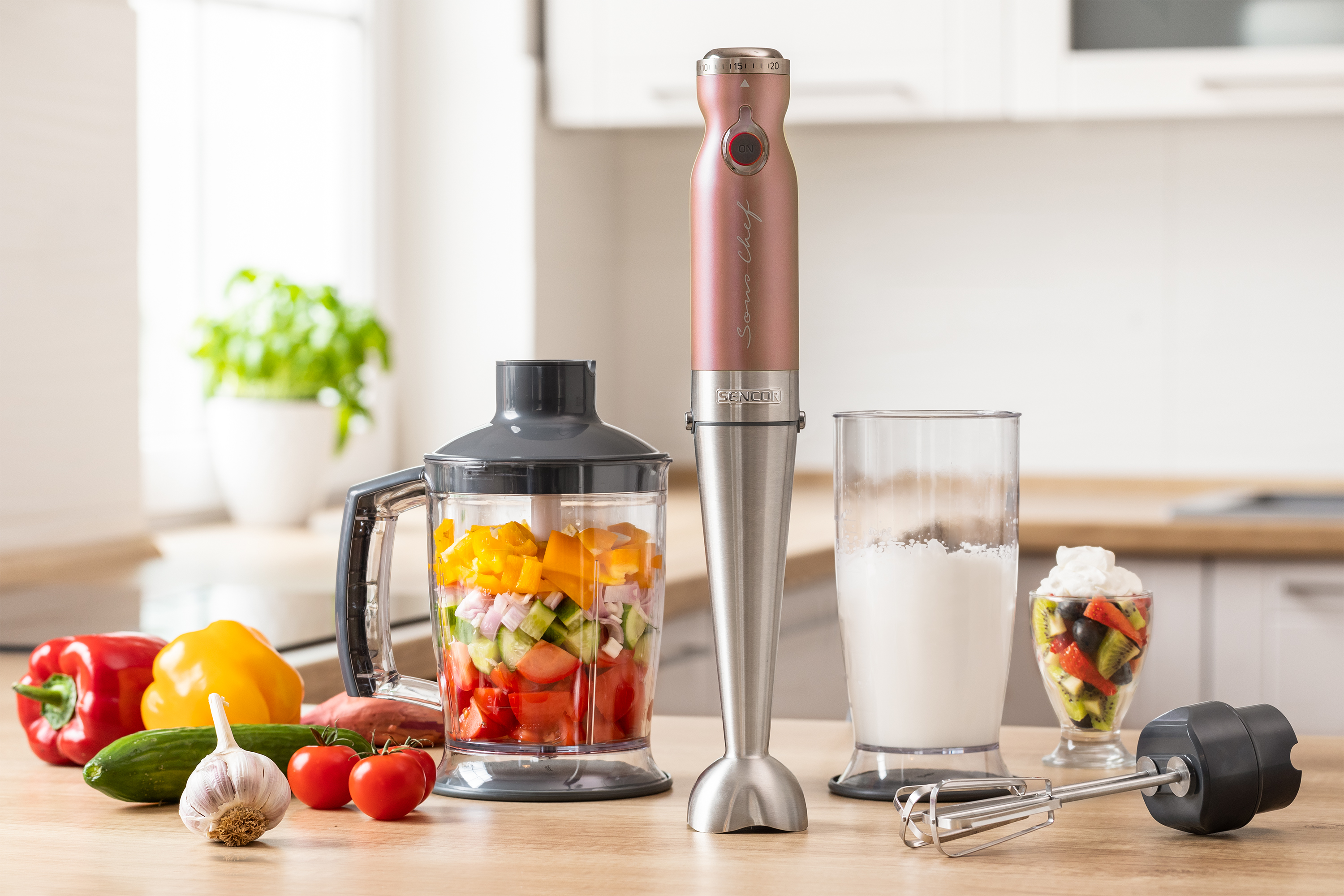 Un frullatore a immersione in oro rosa con accessori e ingredienti su un bancone di legno in un ambiente cucina.