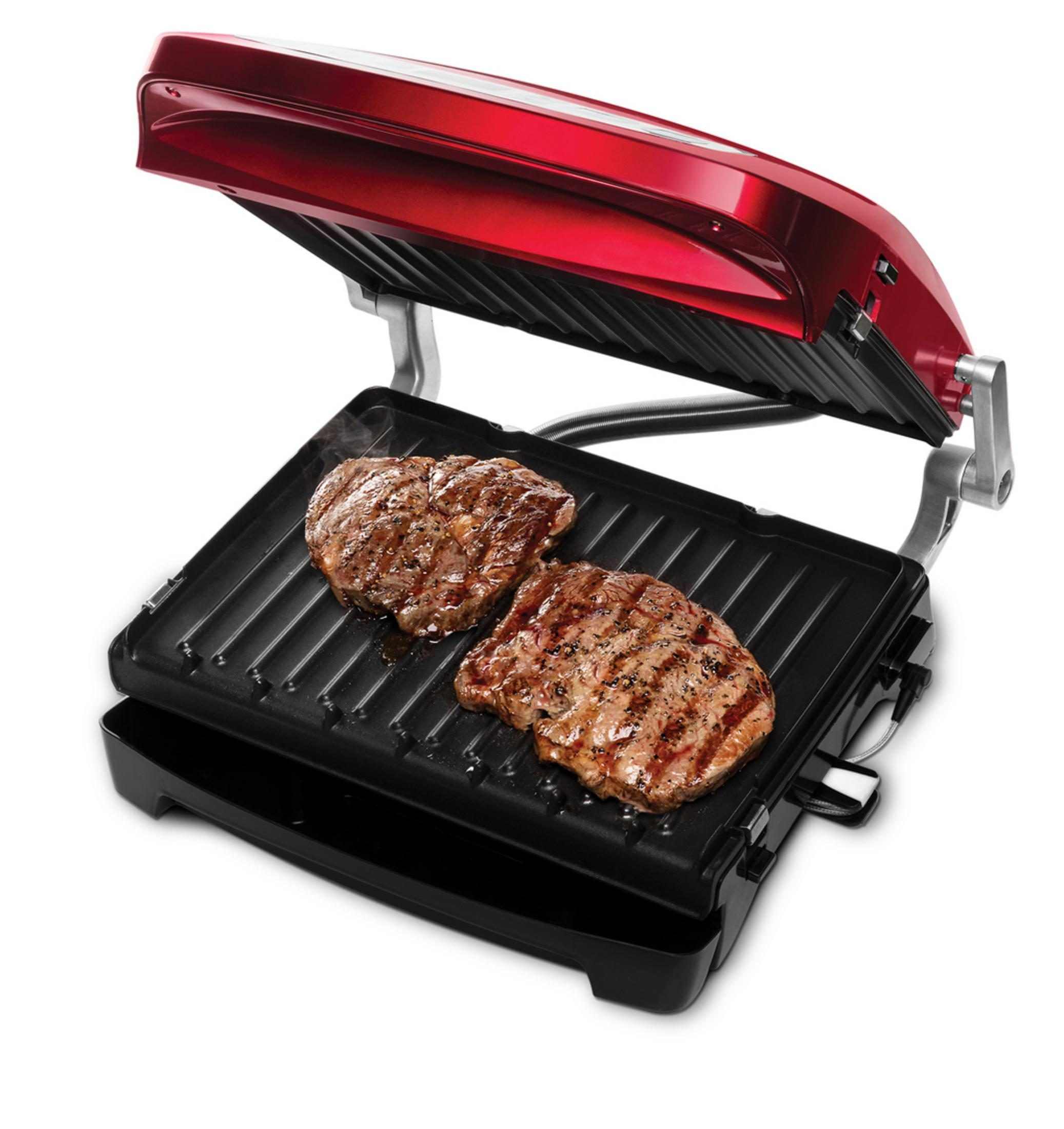 Açık kırmızı bir George Foreman ızgarasında iki biftek ızgara yapılıyor, ızgaranın yapışmaz yüzeyi vurgulanıyor.