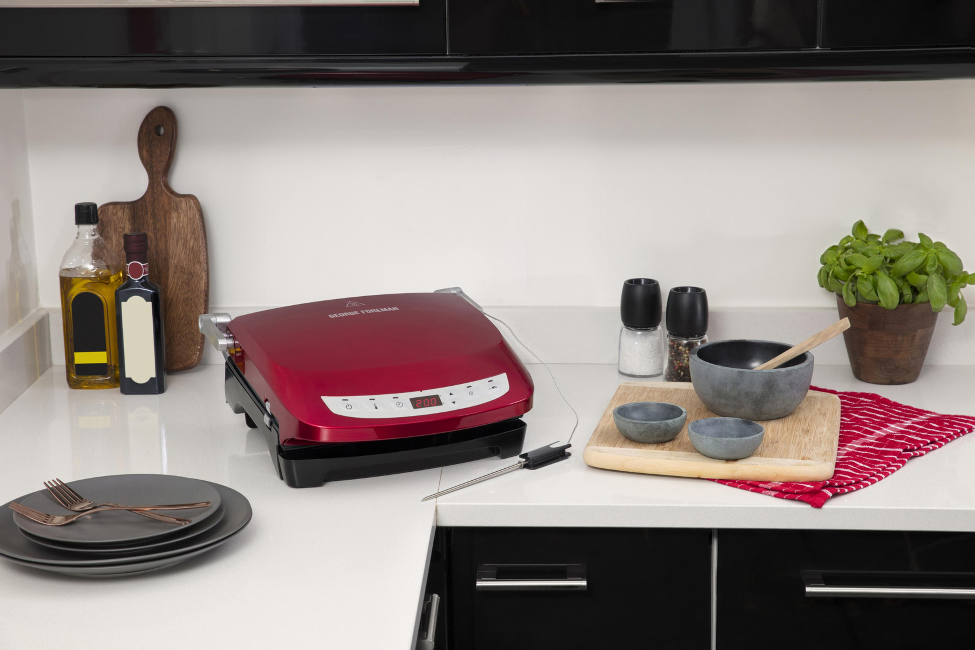Kırmızı bir George Foreman ızgarası, tabaklar, mutfak eşyaları ve yemek pişirme malzemeleriyle çevrili bir mutfak tezgahında duruyor.