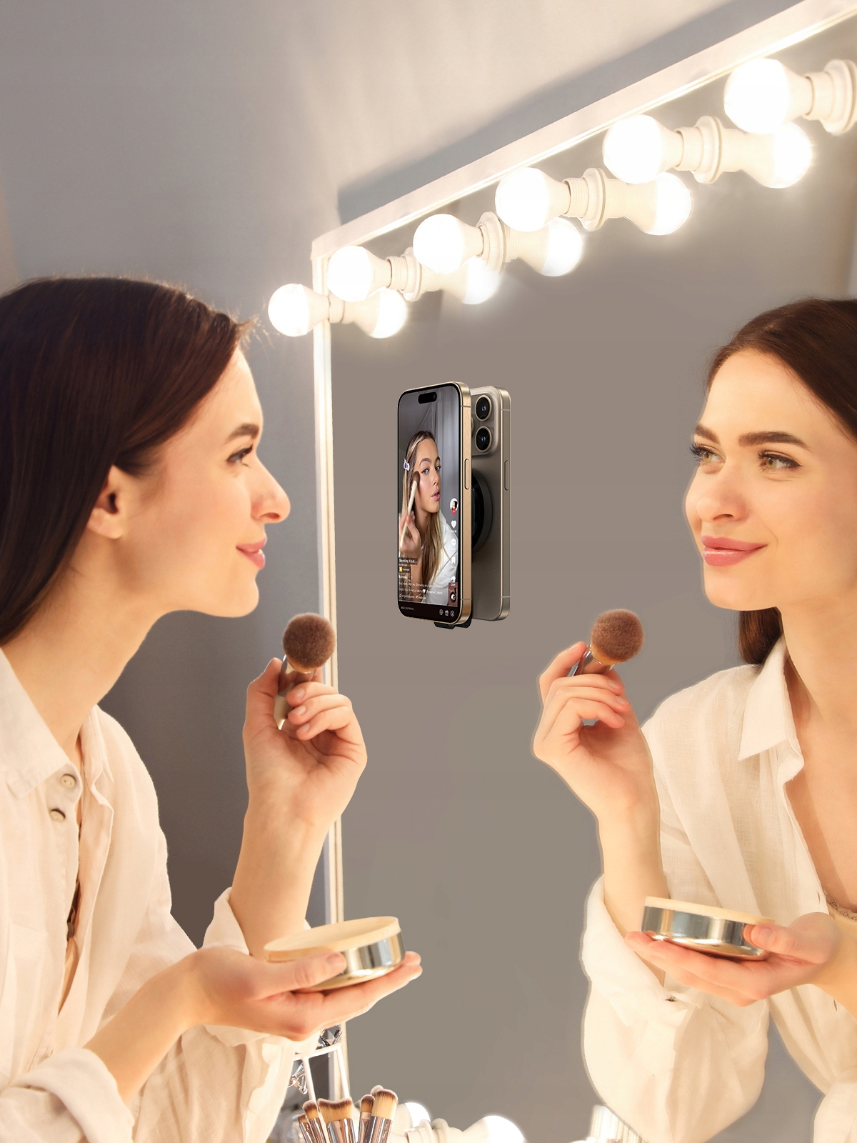 Kobieta nakłada makijaż za pomocą telefonu przymocowanego do lustra za pomocą magnetycznego uchwytu, aby oglądać bez użycia rąk.