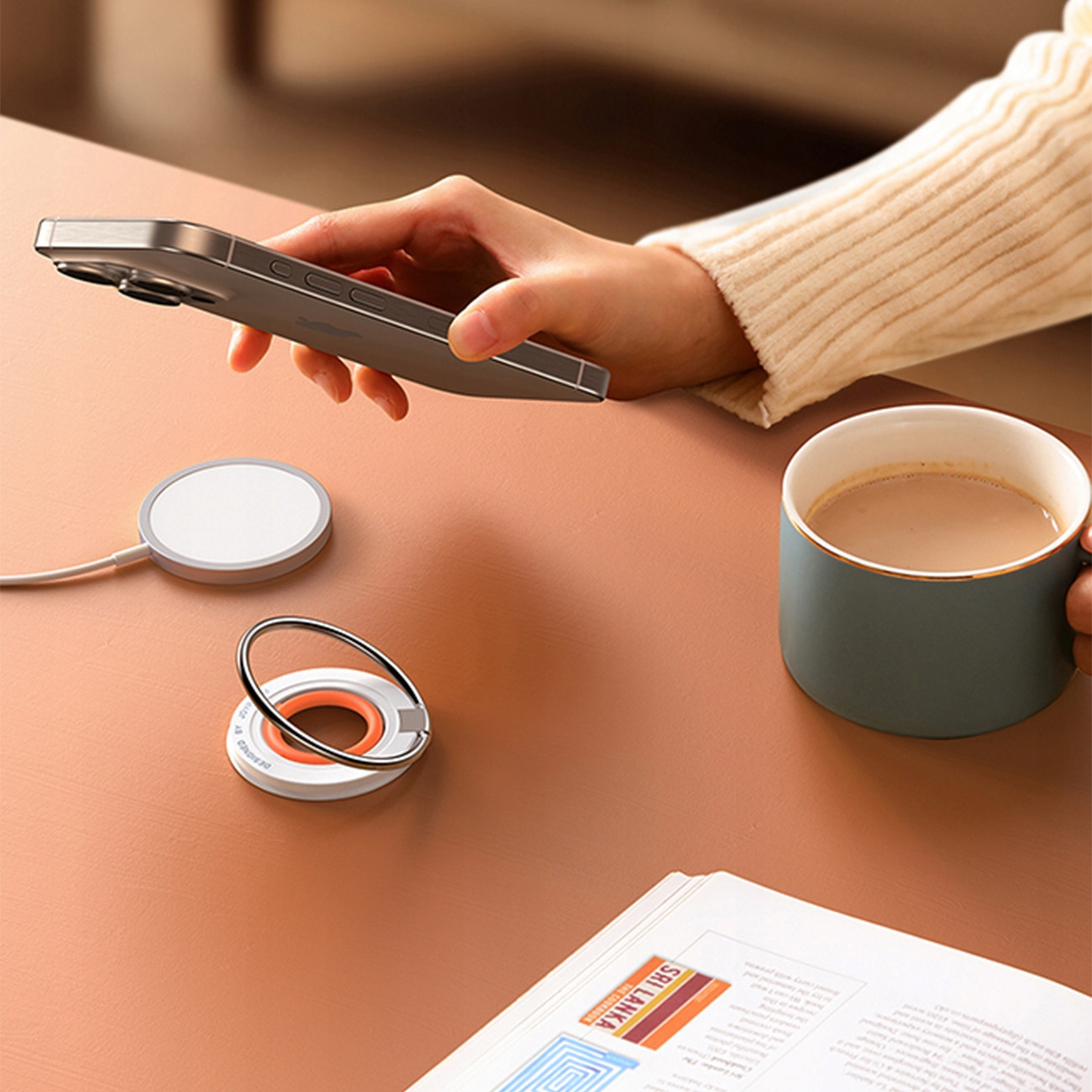 Dłoń trzyma telefon obok ładowarki bezprzewodowej i kubka na biurku. Tło to stół.