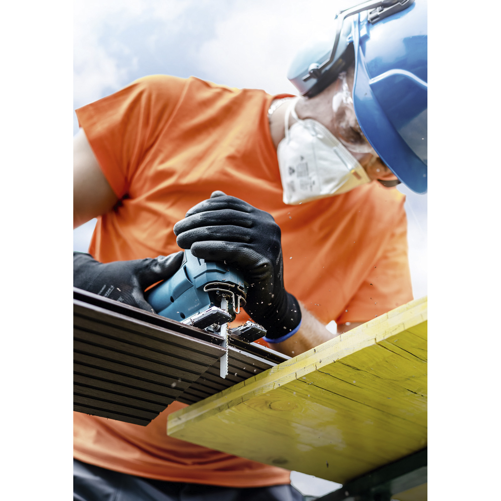 Osoba używająca wyrzynarki, w odzieży ochronnej, tnąca drewnianą deskę na zewnątrz.