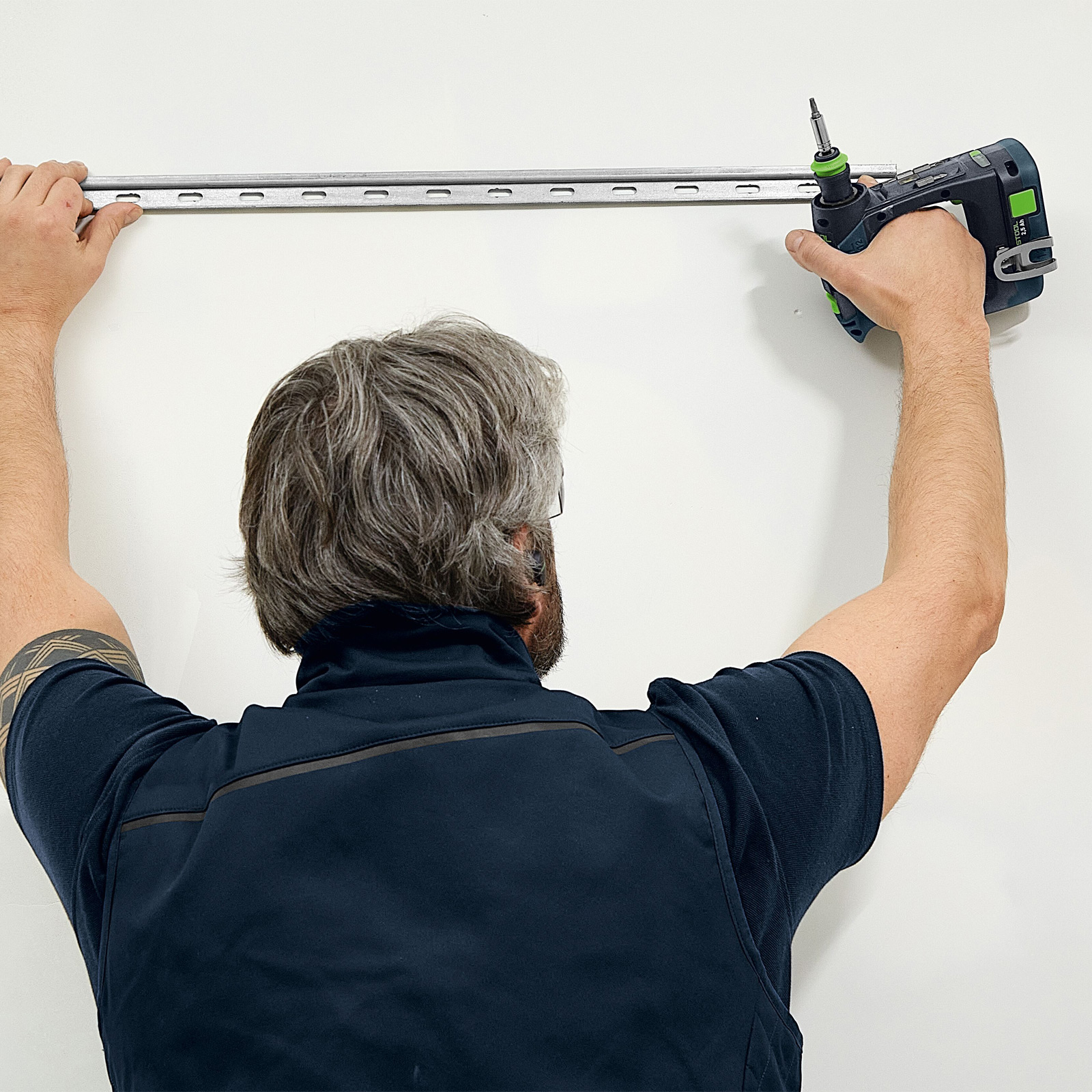 Un hombre instala una guía metálica en una pared blanca con un taladro, de espaldas al espectador.