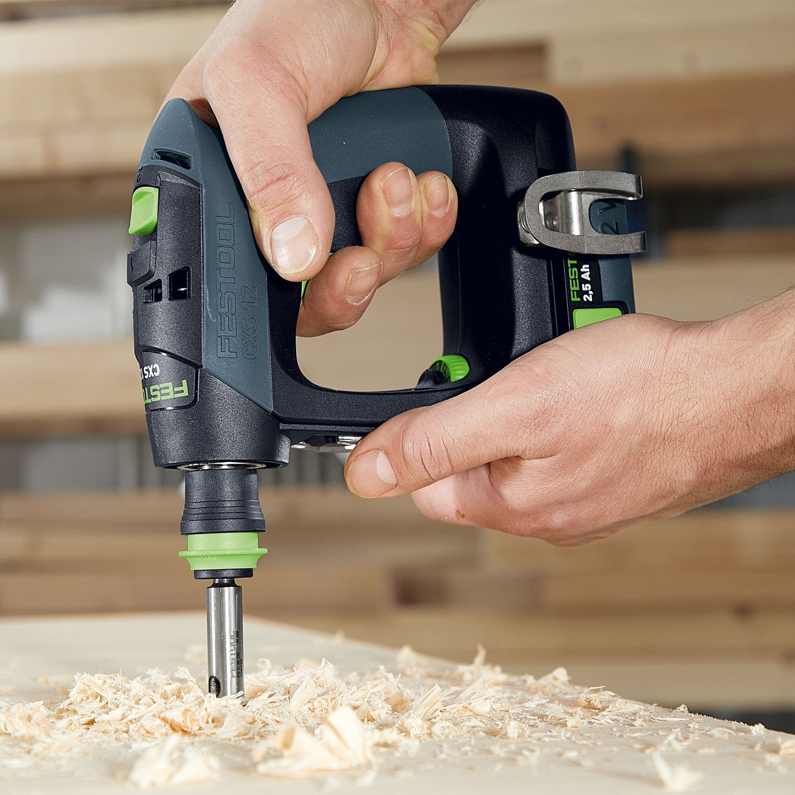 Unas manos sostienen un taladro Festool, perforando madera, rodeado de virutas de madera sobre una superficie de madera.