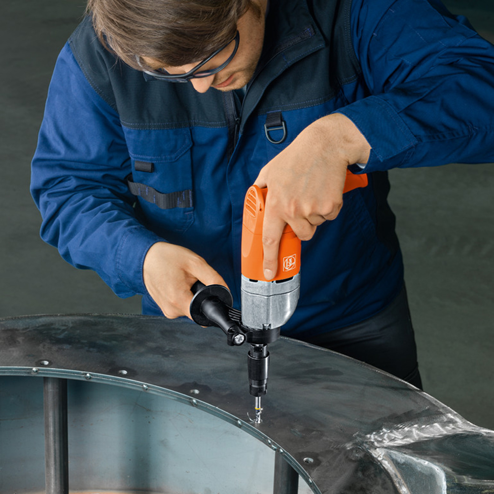 Mann bohrt mit einer orangefarbenen und silbernen Bohrmaschine in eine Metalloberfläche und trägt dabei eine Schutzbrille.