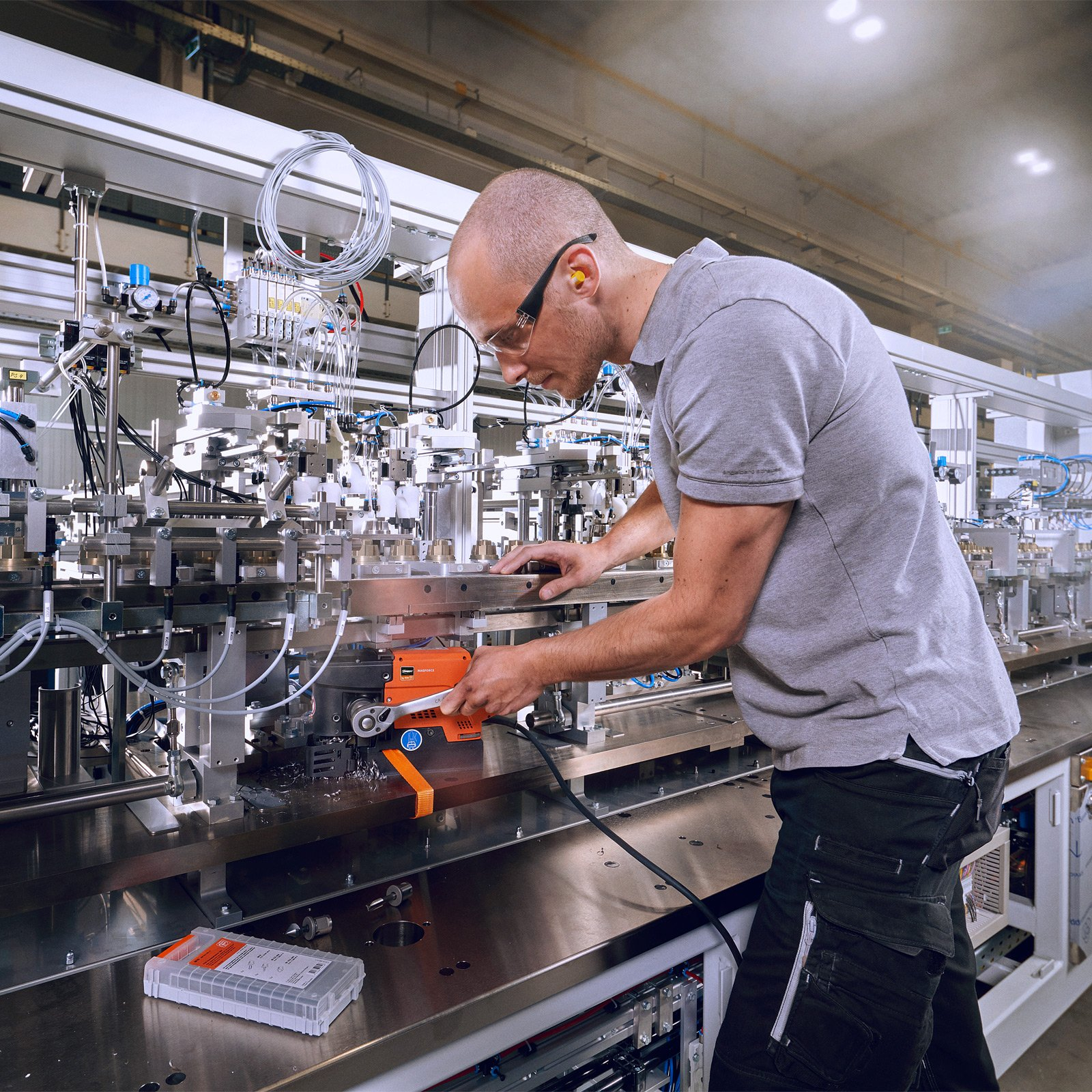 Ein Mann mit Schutzbrille und Ohrstöpseln arbeitet mit einem Schraubenschlüssel an einem industriellen Fließband.