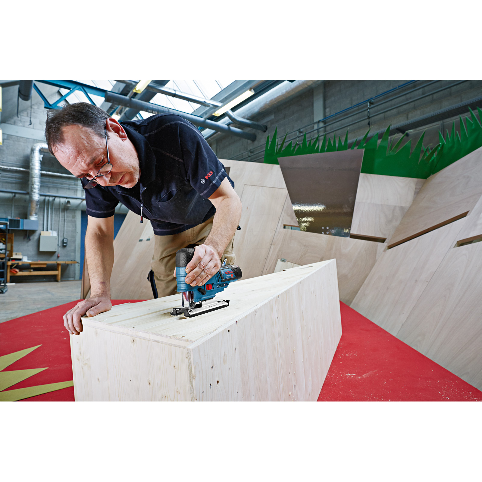 Mężczyzna używa niebieskiej wyrzynarki Bosch do cięcia kawałka drewna w warsztacie.