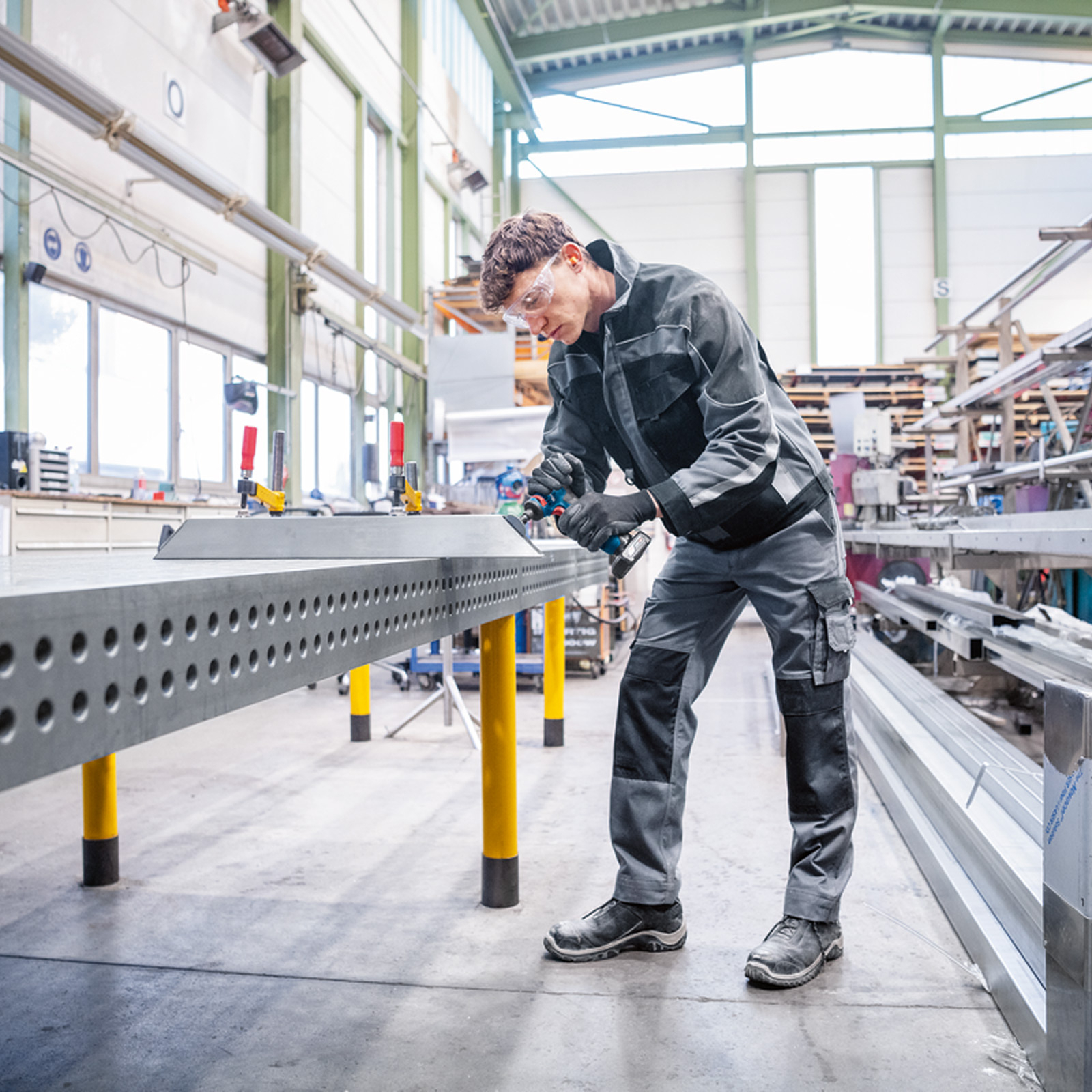 Un travailleur portant des lunettes de sécurité utilise une perceuse sur une table en métal dans un atelier.