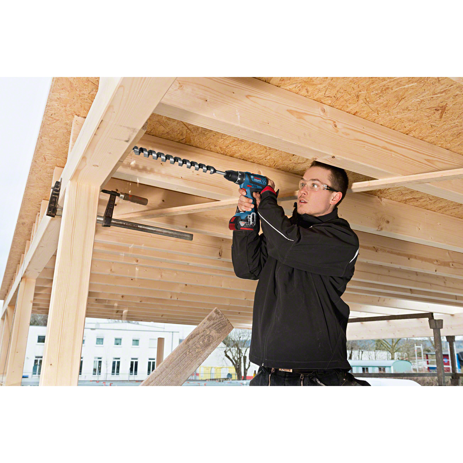 Un hombre taladra en una estructura de madera con un taladro inalámbrico Bosch azul en una obra.