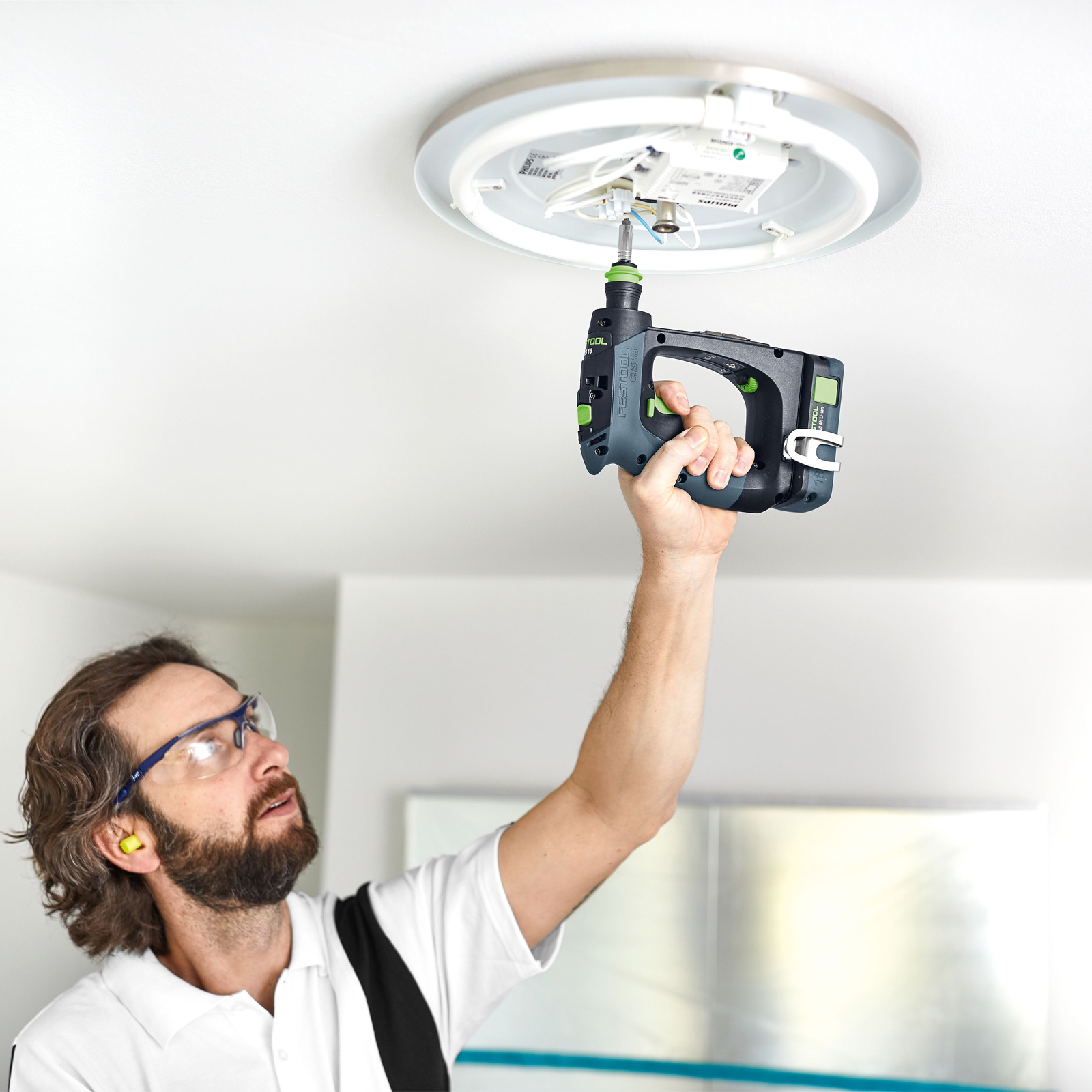 Un uomo installa una plafoniera con un trapano Festool, indossando occhiali di sicurezza e tappi per le orecchie.