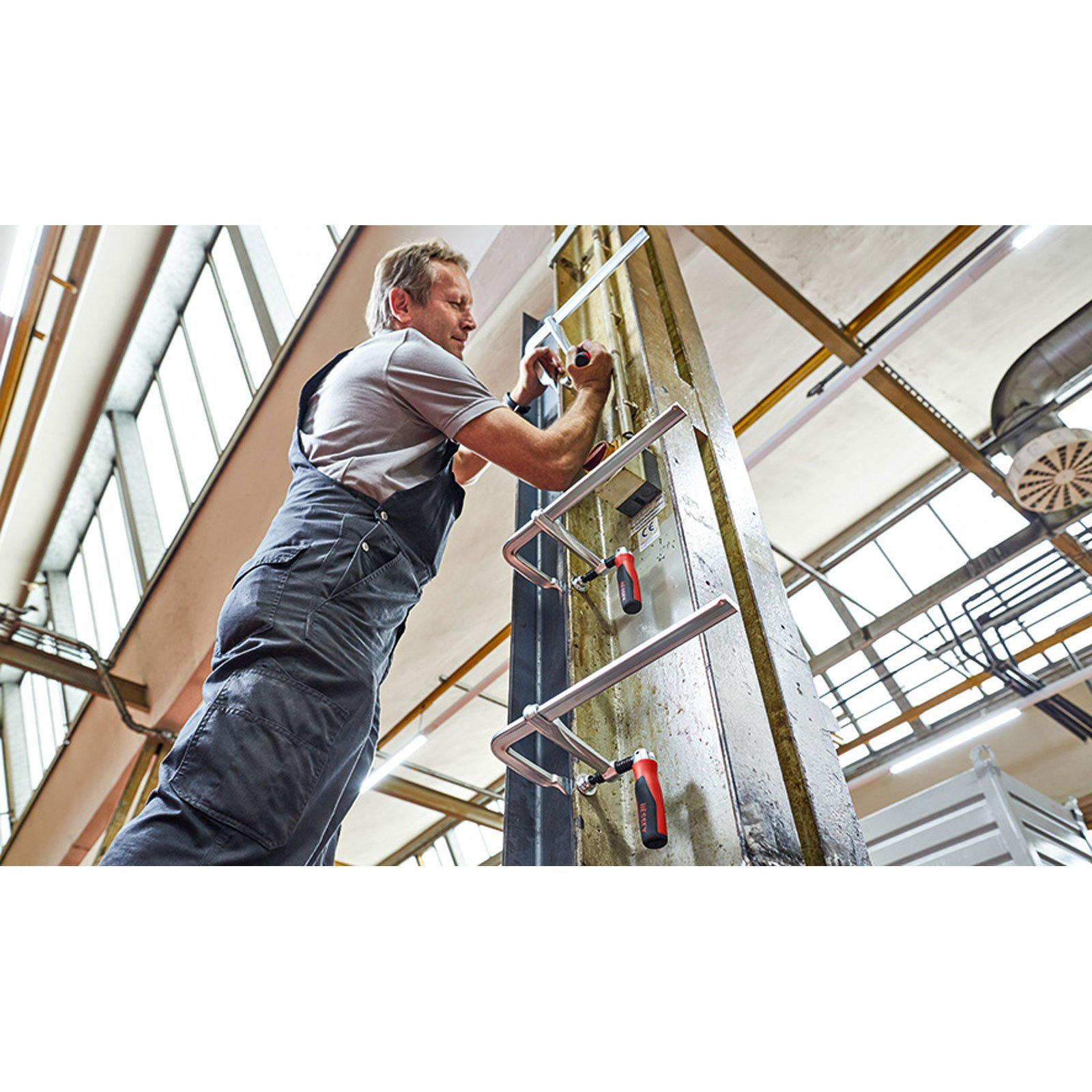 Mężczyzna pracuje z zaciskami na konstrukcji. Ma na sobie kombinezon i stoi w warsztacie.