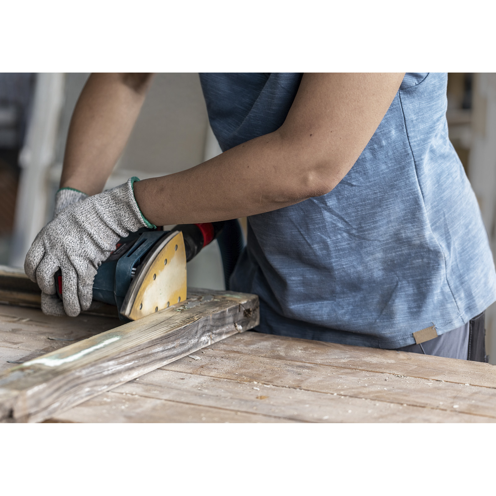 Person schleift Holz mit einem Elektroschleifer und trägt dabei Handschuhe. Die Szene spielt in einer Werkstatt.