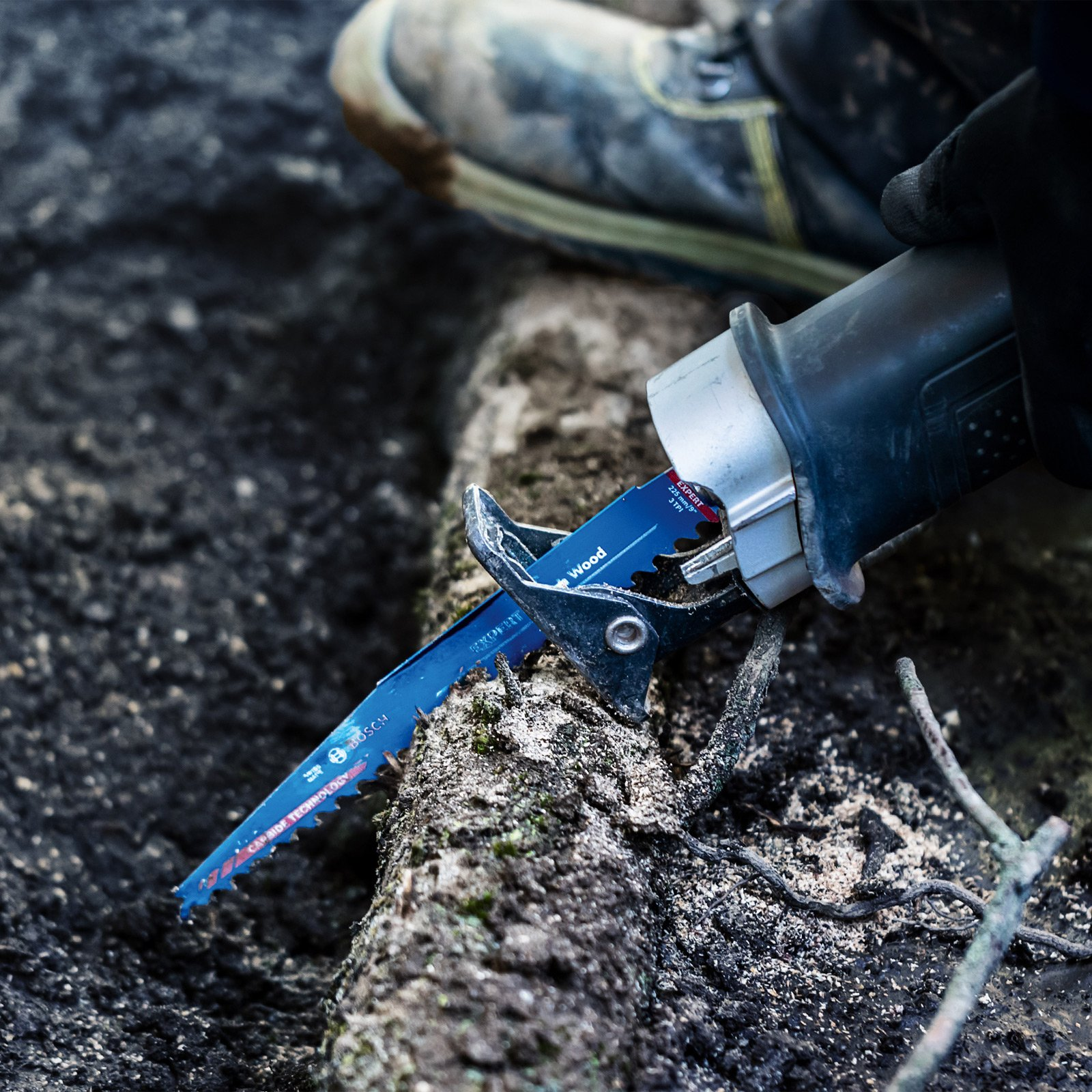 Zbliżenie niebieskiego ostrza piły przecinającego drewno w pobliżu buta na kamienistej powierzchni.