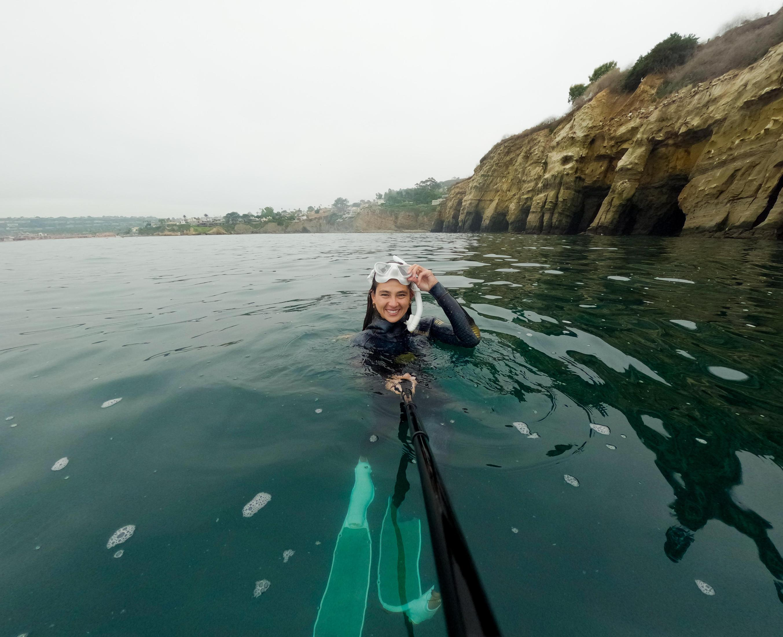 Una donna nell'oceano che tiene una GoPro su un bastone, con delle scogliere dietro.