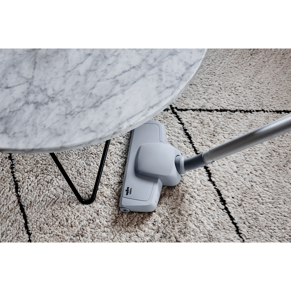Tête d'aspirateur grise sous une table en marbre sur un tapis blanc avec des lignes noires.